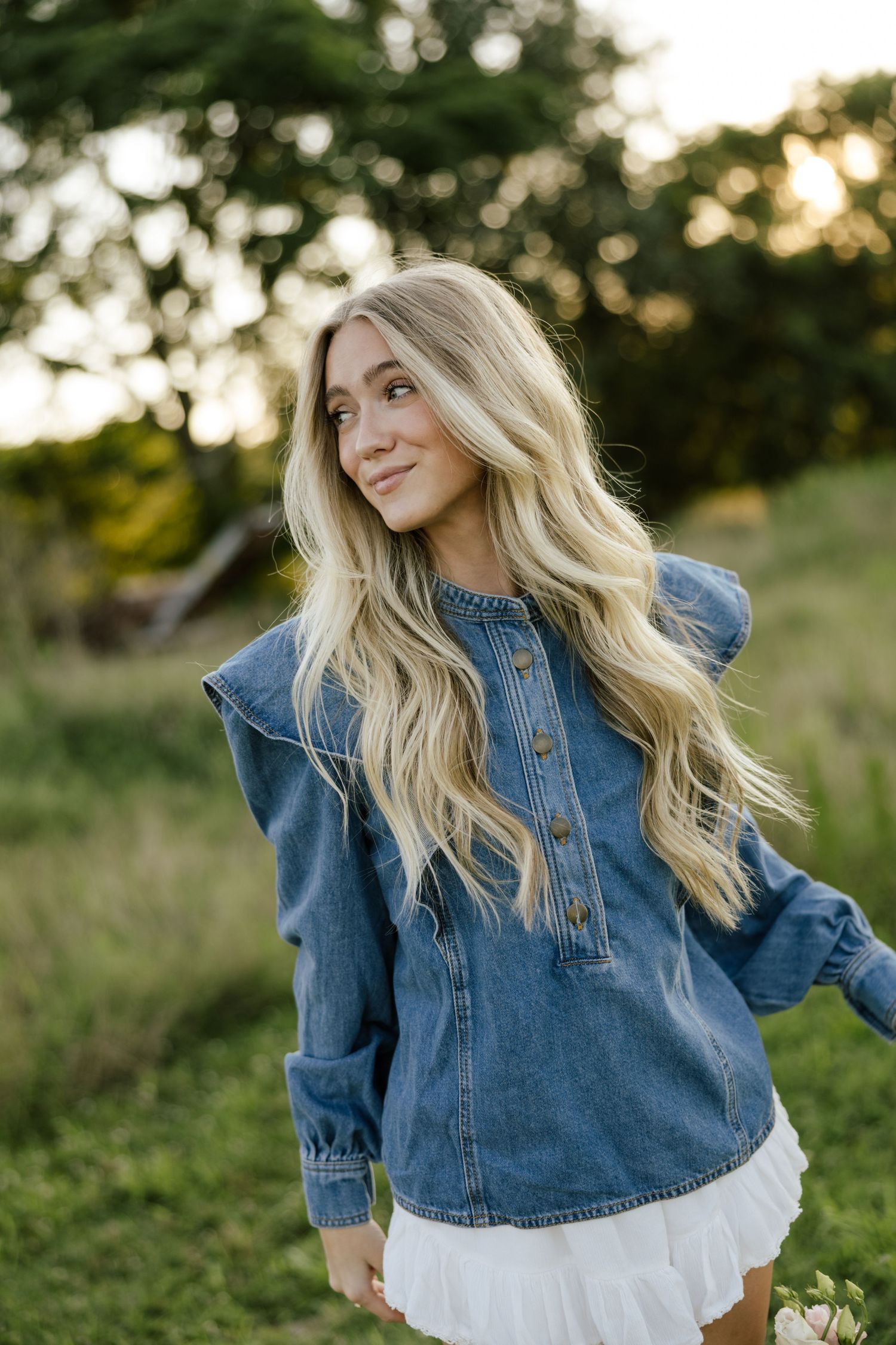 Blonde woman in denim ruffle jacket and white skirt posing outdoors in a green field at golden hour.