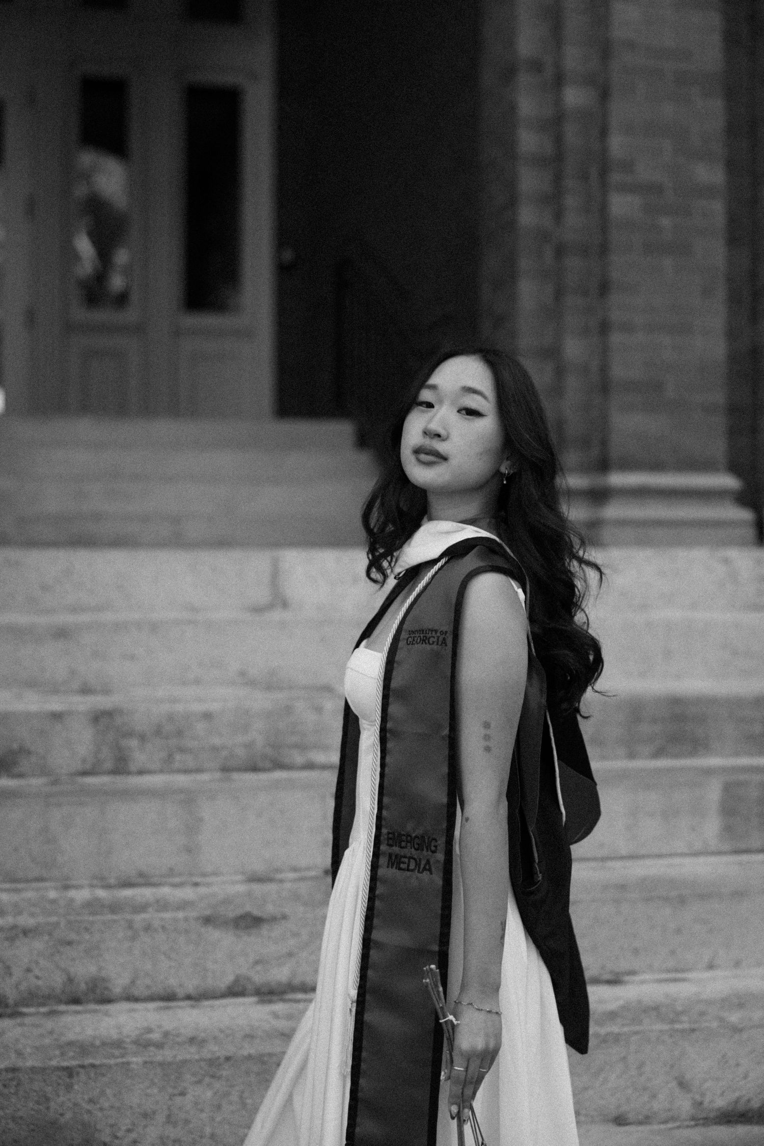 Black and white photo of a young woman in graduation attire with sash, posing on steps outside a building.