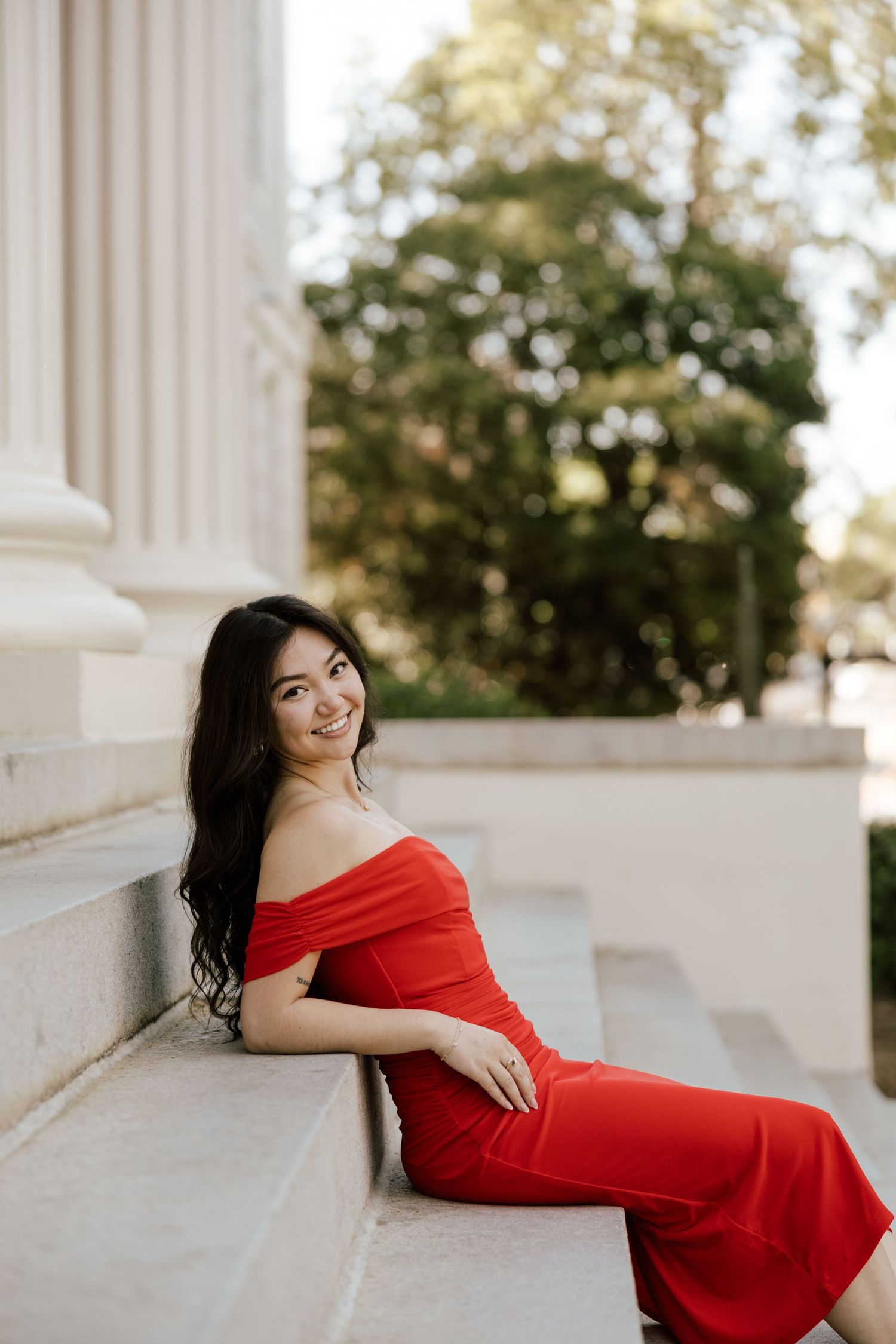 Young woman in red off-shoulder gown sitting gracefully on marble steps beside white columns outdoors.