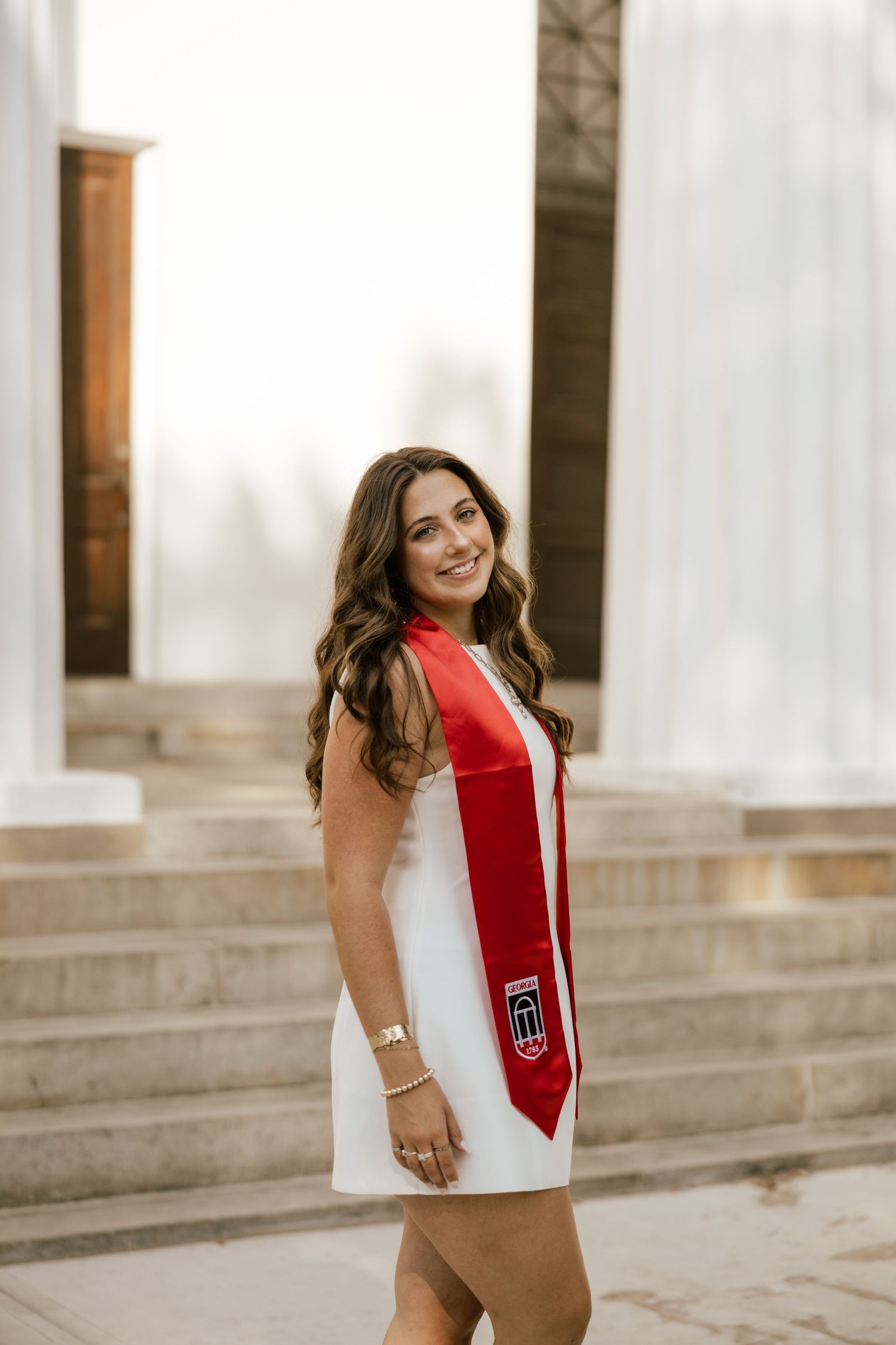 Young woman in white dress and red graduation stole smiles outdoors near stone columns and steps.