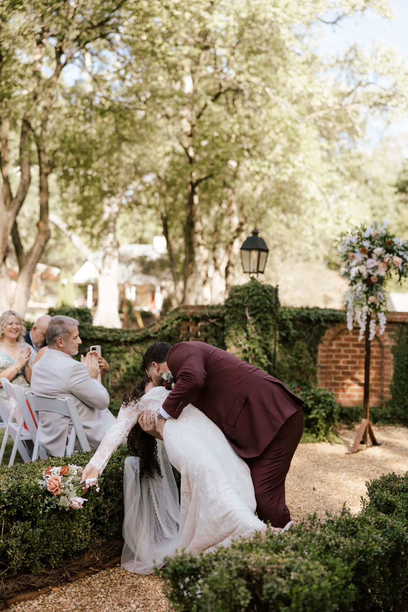 Groom in burgundy suit dips bride in white gown for a romantic kiss at an outdoor garden wedding ceremony.