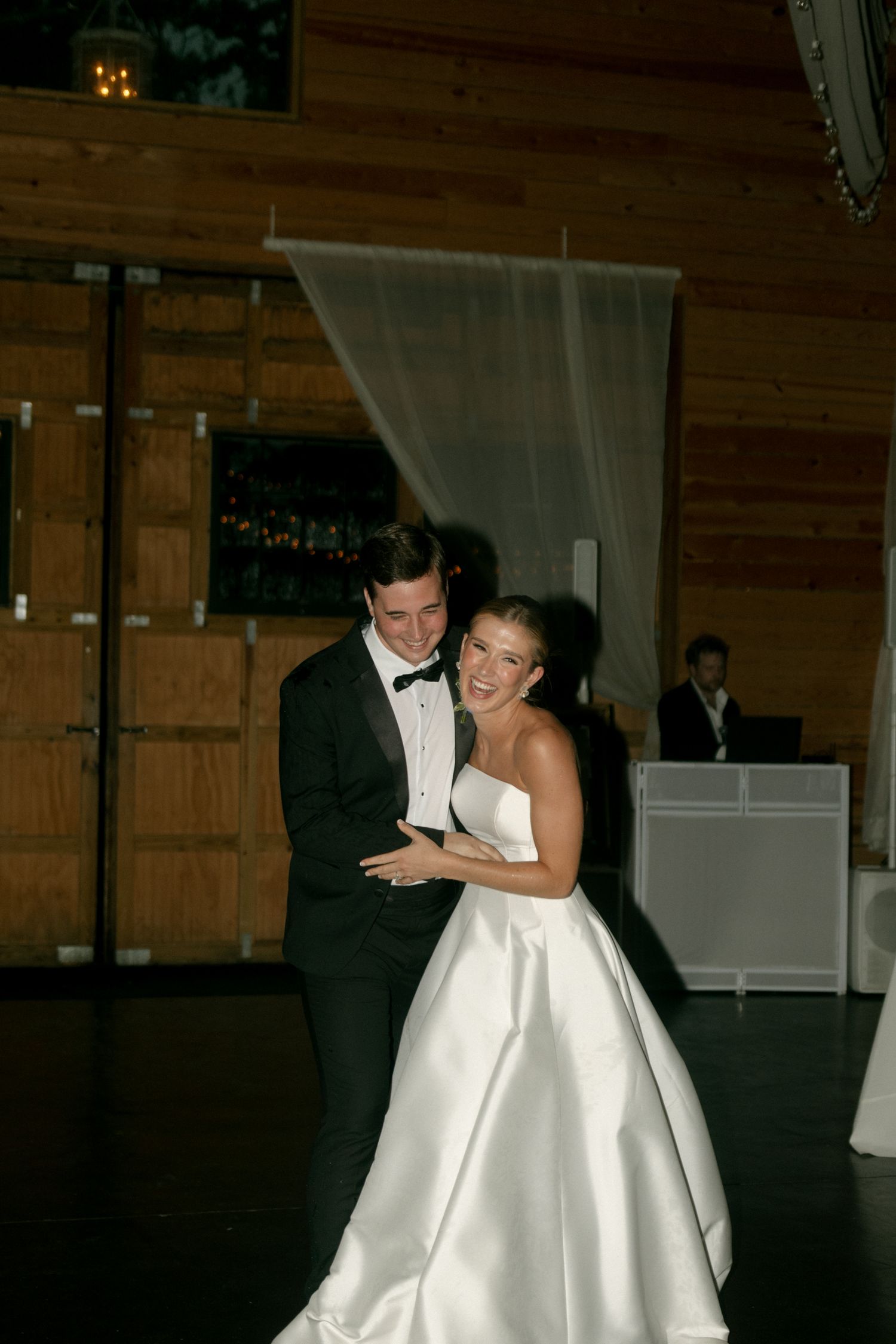 Bride and groom share their first dance, smiling joyfully in a rustic wood-paneled wedding venue.