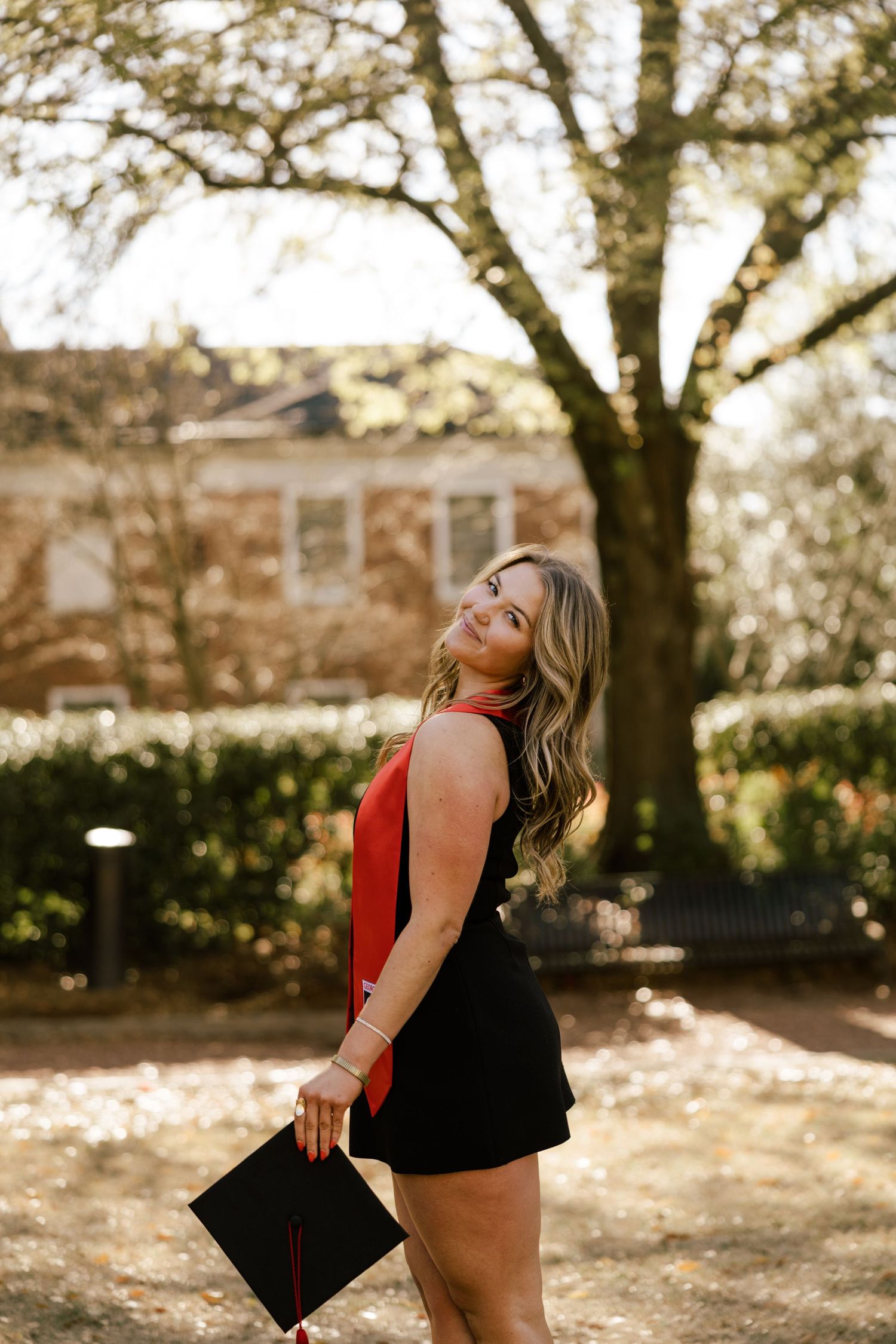 A person in a red top and black shorts laughs joyfully while standing under autumn trees in a park setting.
