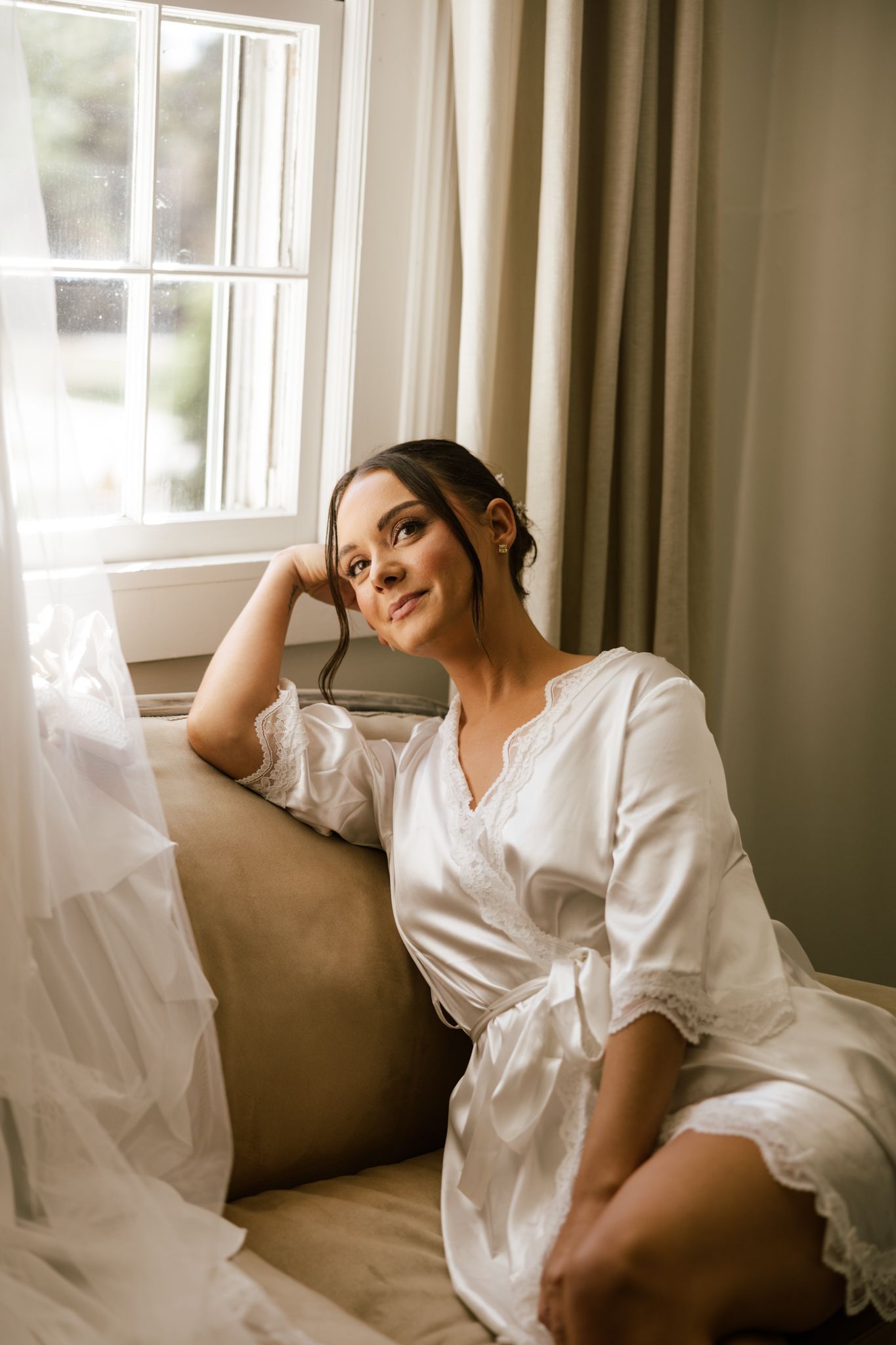 Bride in white lace robe sitting by window on wedding morning, gazing upward in soft natural light.