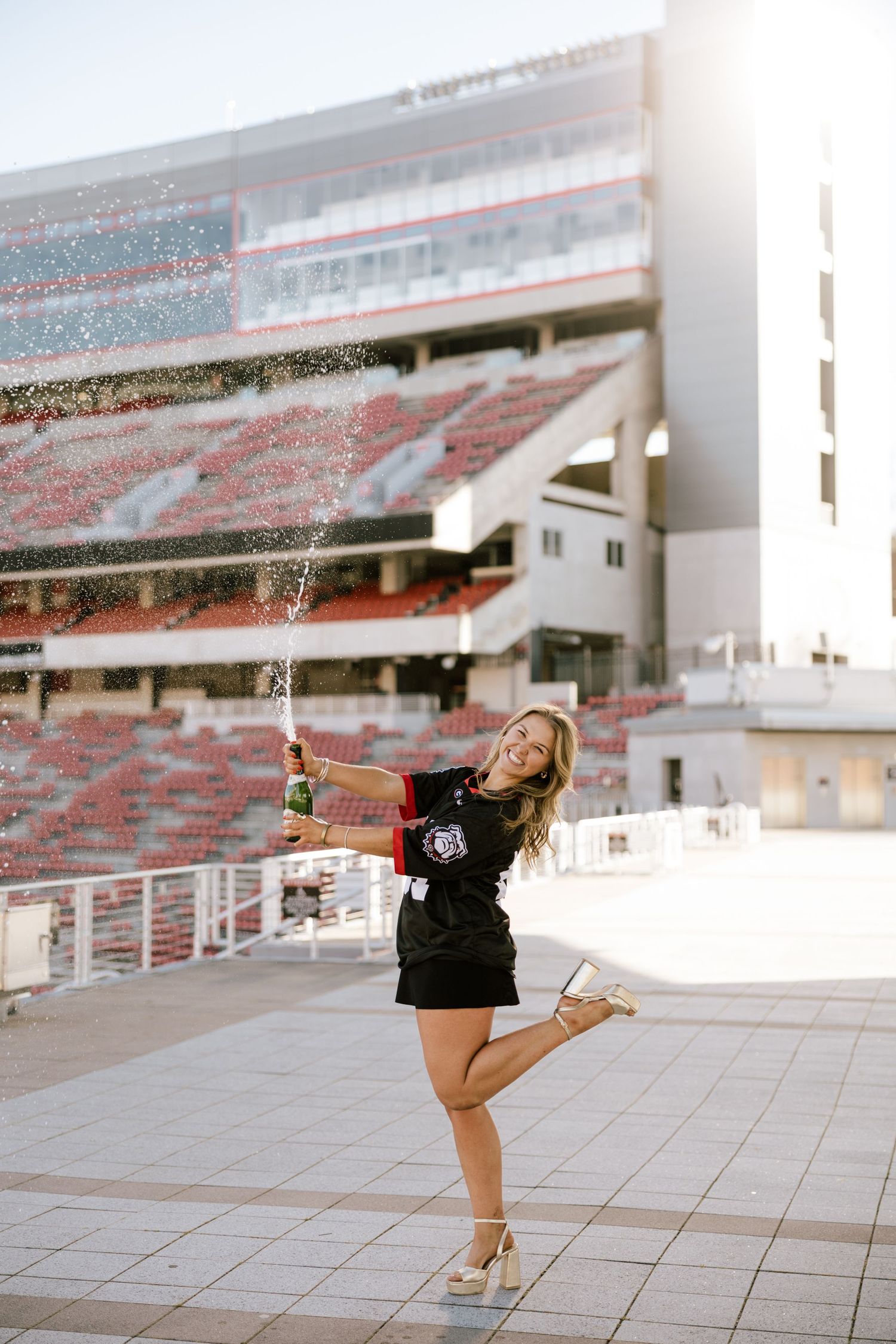 Woman in black jersey celebrates with champagne spray at a large college football stadium on a sunny day.
