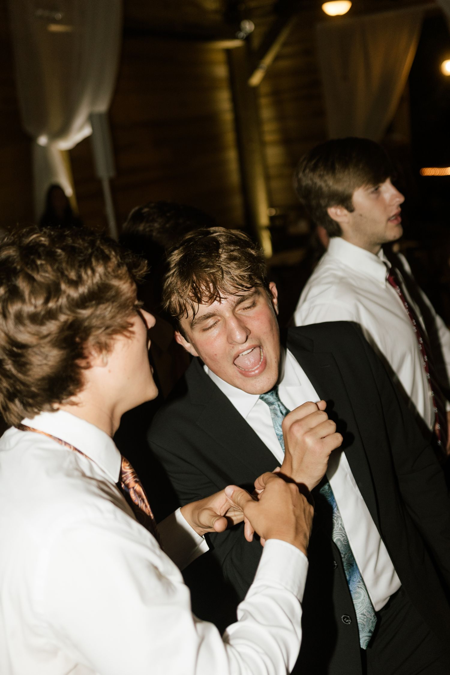 Young men in suits and ties dancing energetically at a formal event or wedding reception.