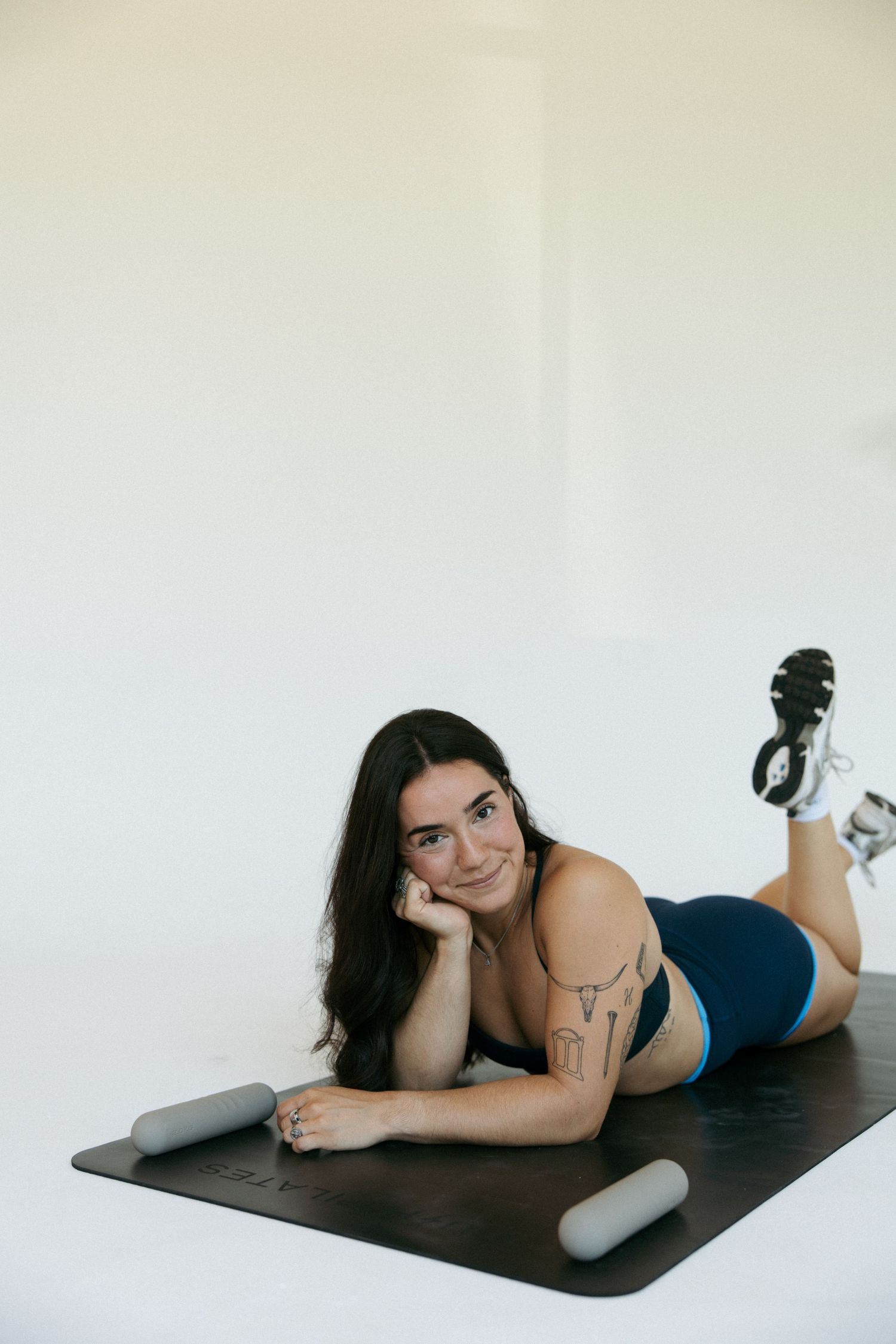 Woman in blue activewear lying on yoga mat with foam rollers, posing thoughtfully in a bright white studio.