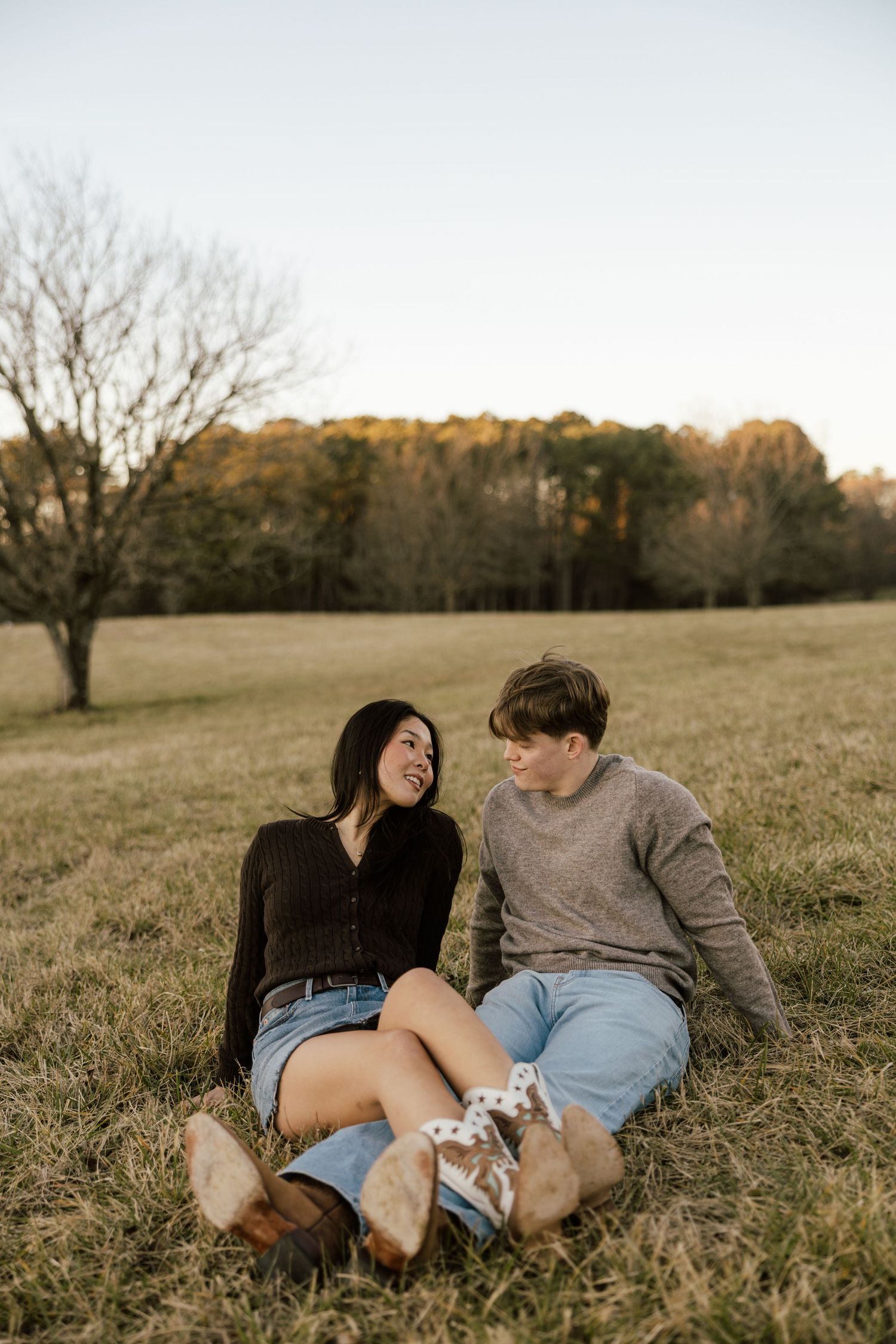 A young couple sits together on a grassy field, smiling at each other during golden hour.