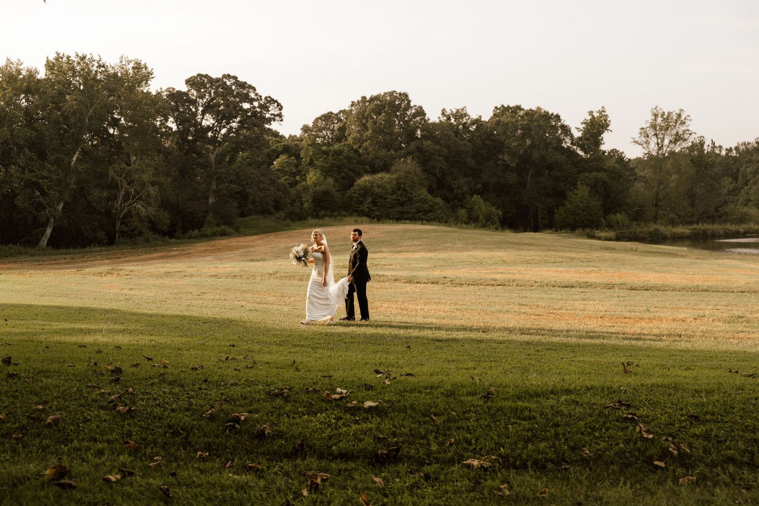 Bride and groom stand together on a lush green golf course at sunset surrounded by trees.