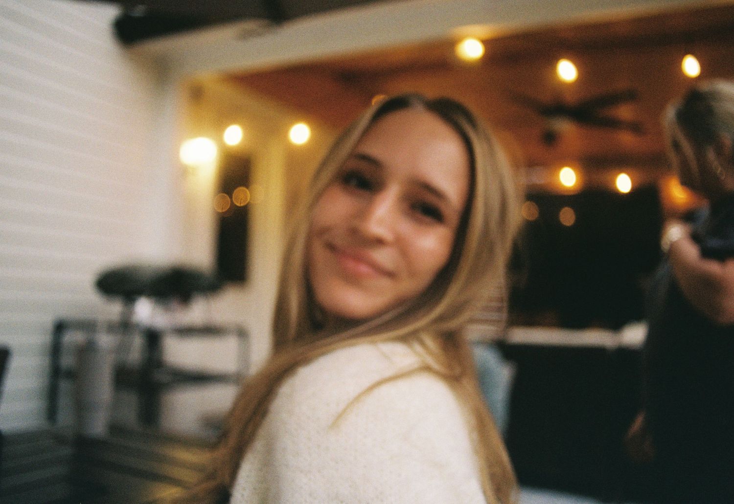 A person smiles while sitting in a dimly lit restaurant with warm ambient lighting in the background.