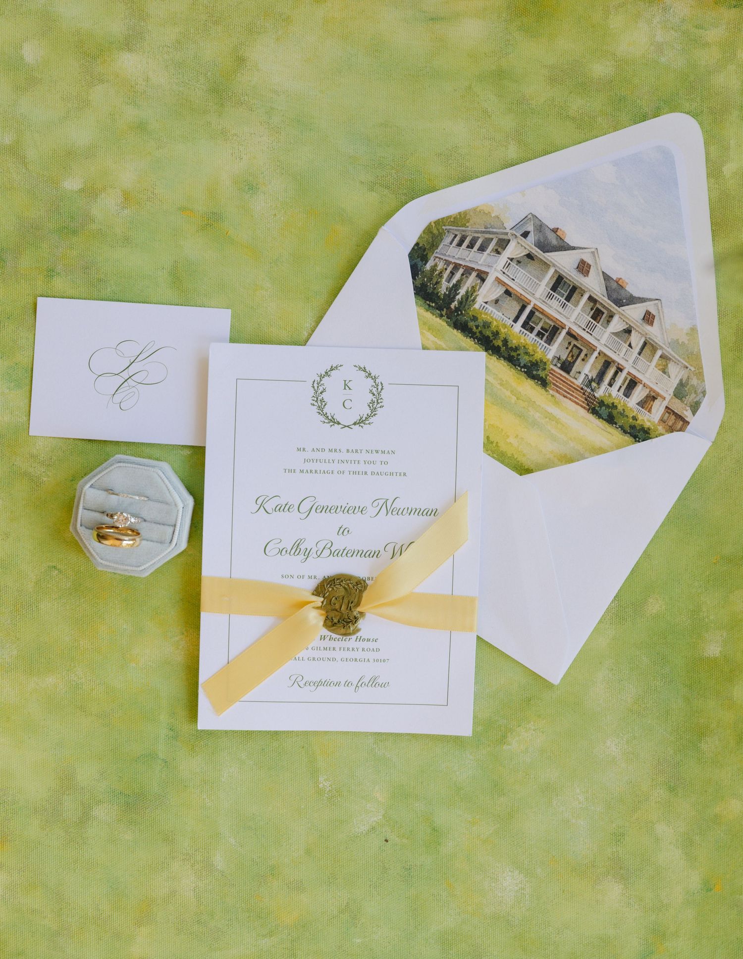 Elegant wedding invitation suite with yellow ribbon, wax seal, watercolor venue envelope liner, and ring box on green background.