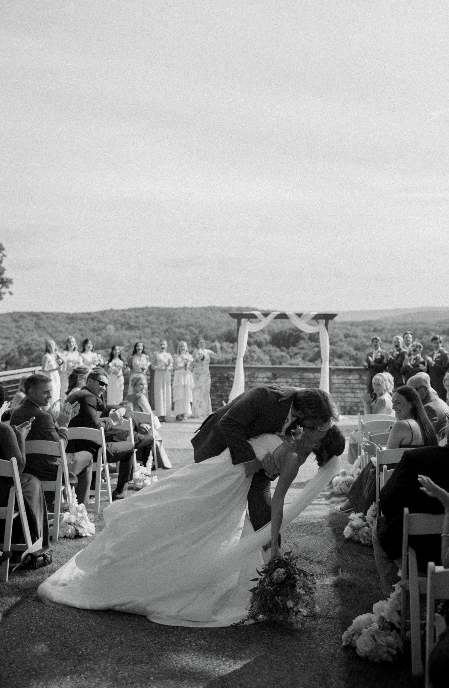 Black and white photo of bride and groom sharing a romantic dip kiss at outdoor wedding ceremony with guests applauding.