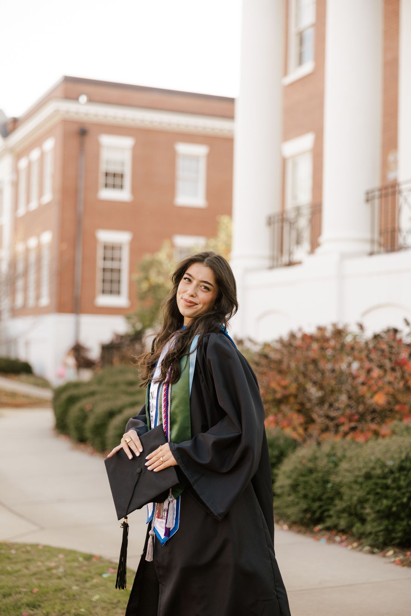 Young woman in black graduation gown and cap poses outdoors near brick university building on campus.
