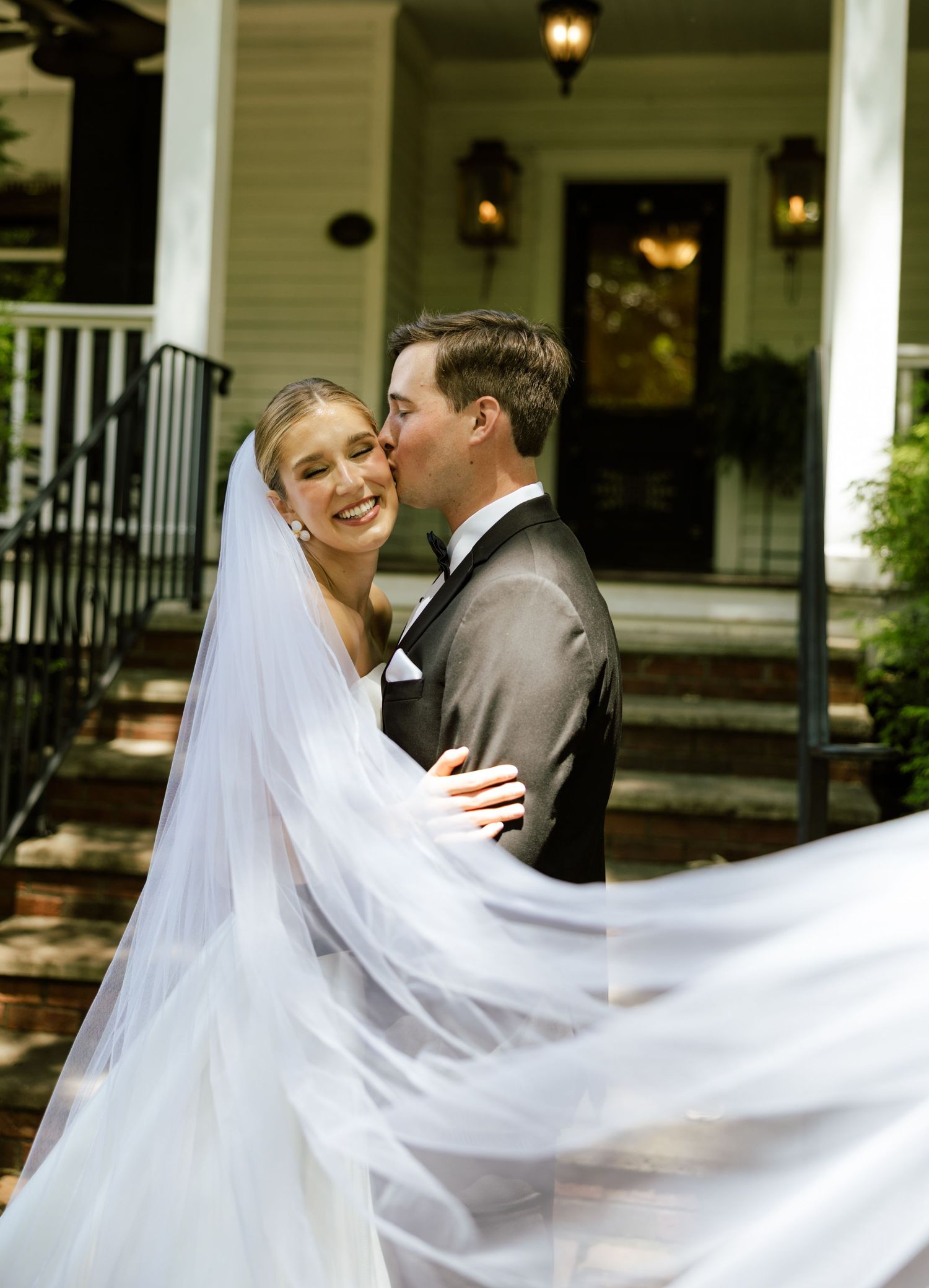A bride in a flowing veil and groom in a tuxedo share a romantic kiss on steps of a white house.