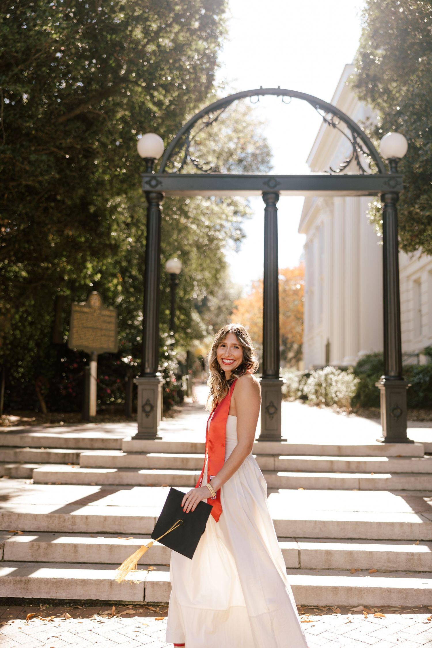 Outdoor photoshoot featuring white dress and red sash against architectural columns and steps.
