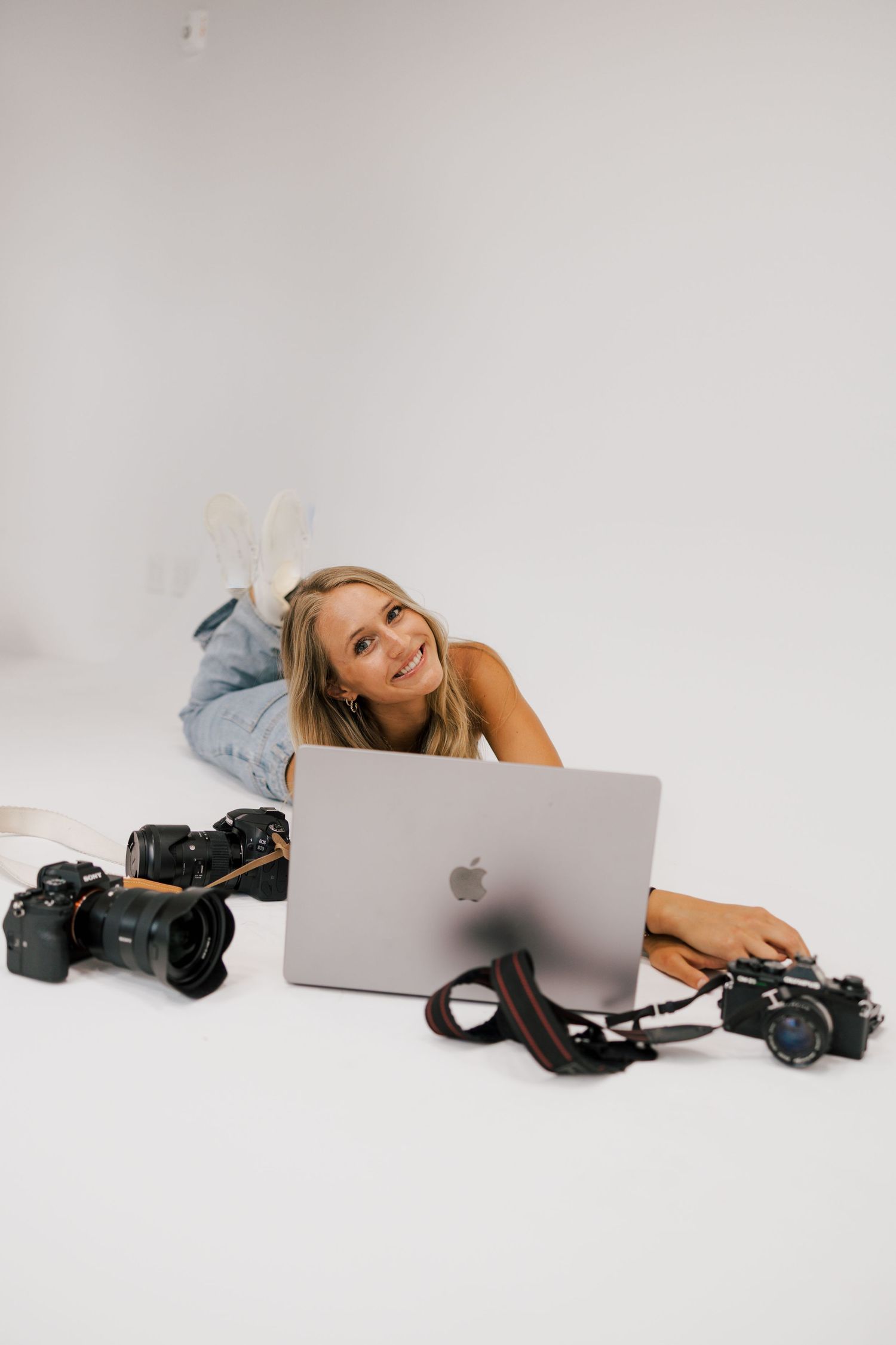 A photographer lies on white backdrop with laptop and camera equipment.