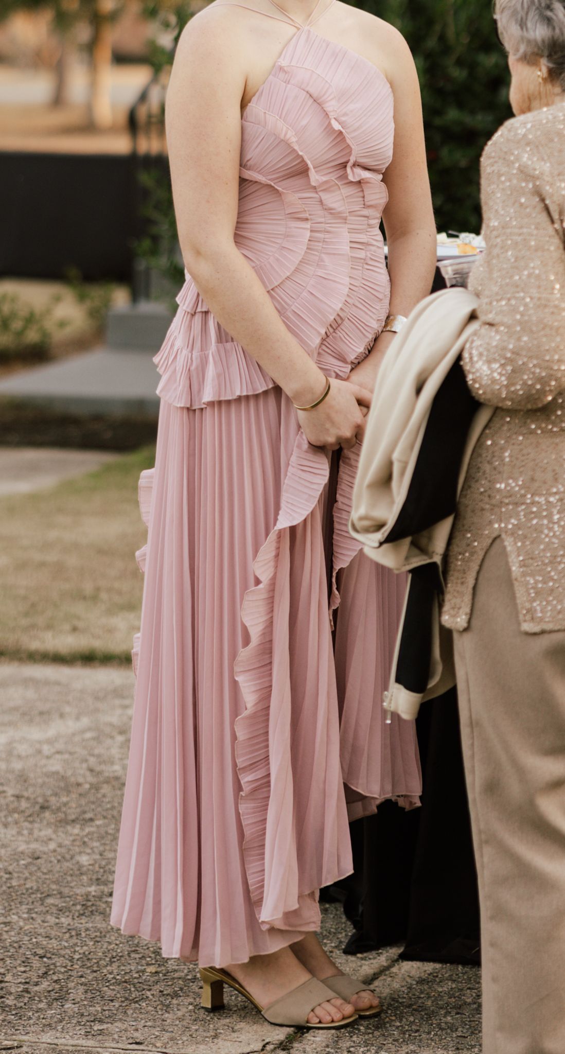 Elegant blush pink pleated ruffle midi dress with halter neckline worn at a formal outdoor event.