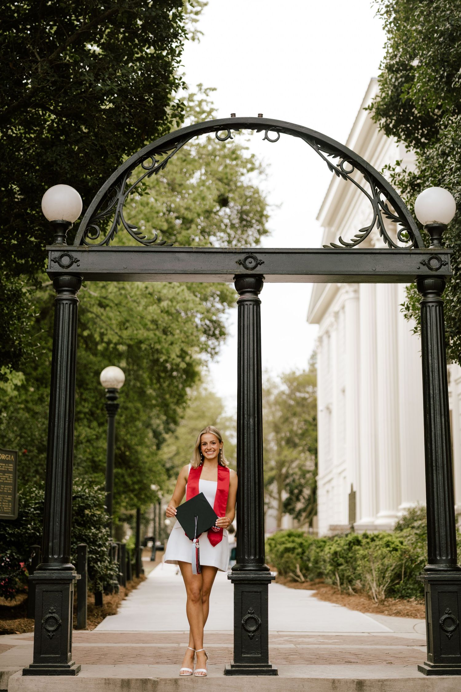 A smiling graduate in a white dress and red jacket poses under an ornate black arch on a college campus.