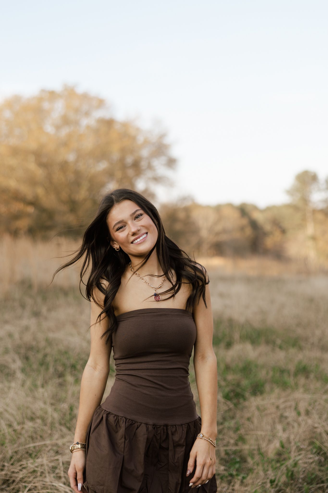 A smiling young woman with dark hair wearing a brown tube top and skirt stands in a golden field at sunset.