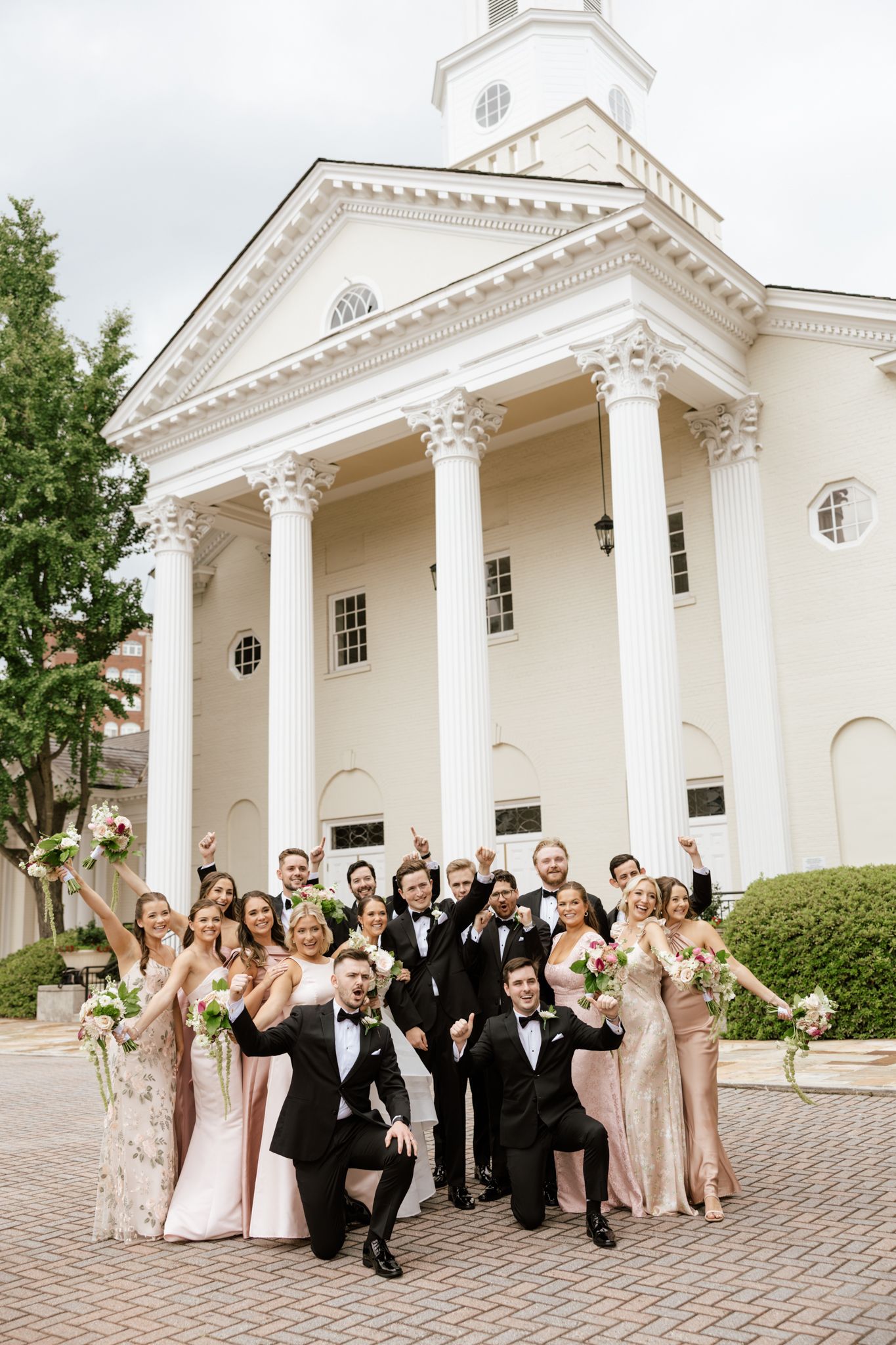 Wedding party celebrating in front of a white colonial church with columns, bridesmaids in pink gowns and groomsmen in black tuxedos.