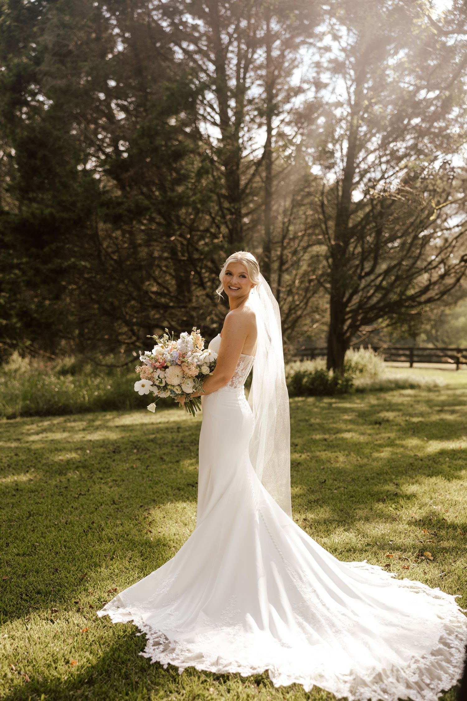 Bride in white mermaid gown with lace train and veil holds bouquet outdoors in sunlit garden.