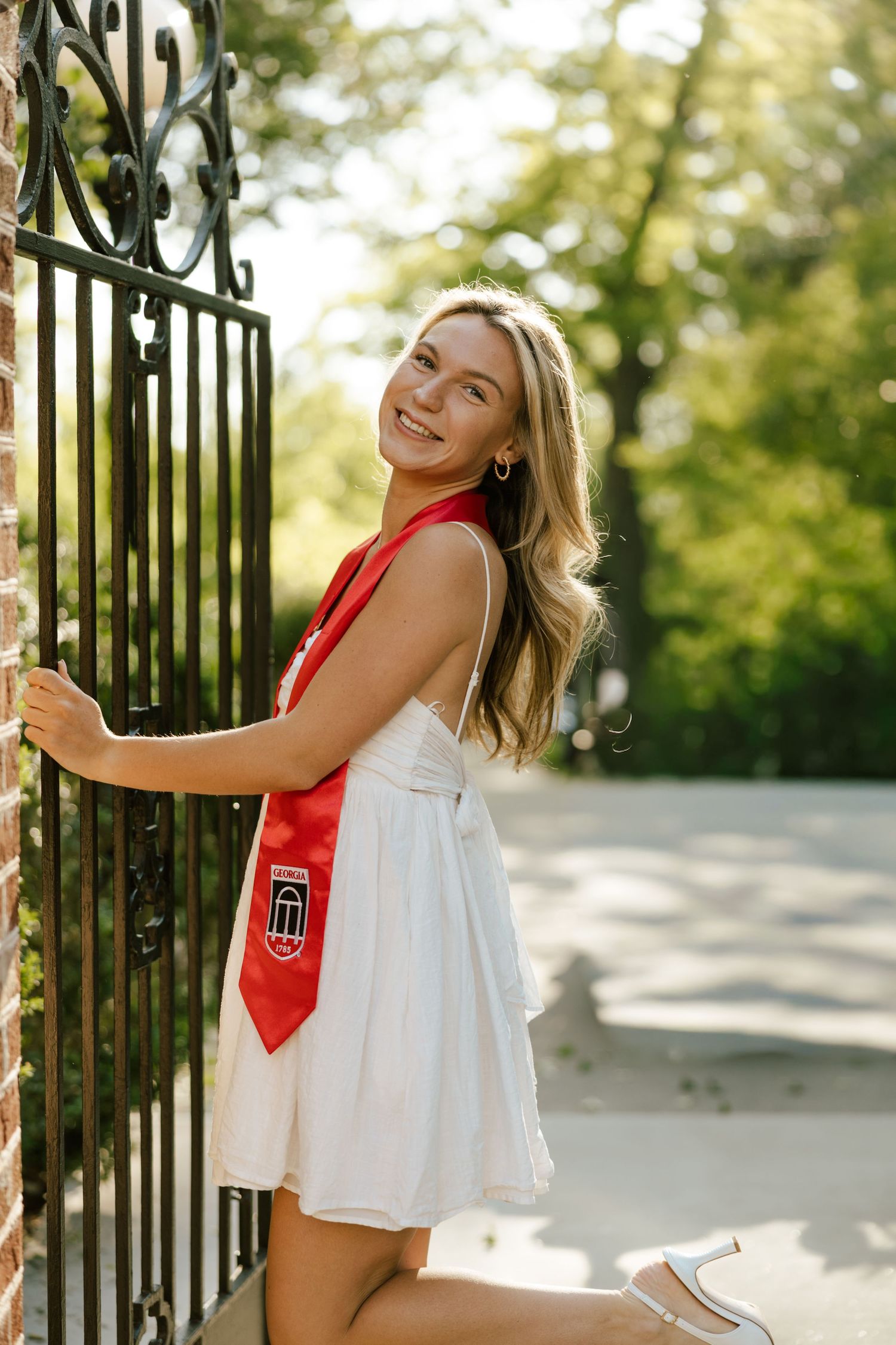 Smiling blonde woman in white dress and red graduation sash poses by ornate iron gate outdoors.