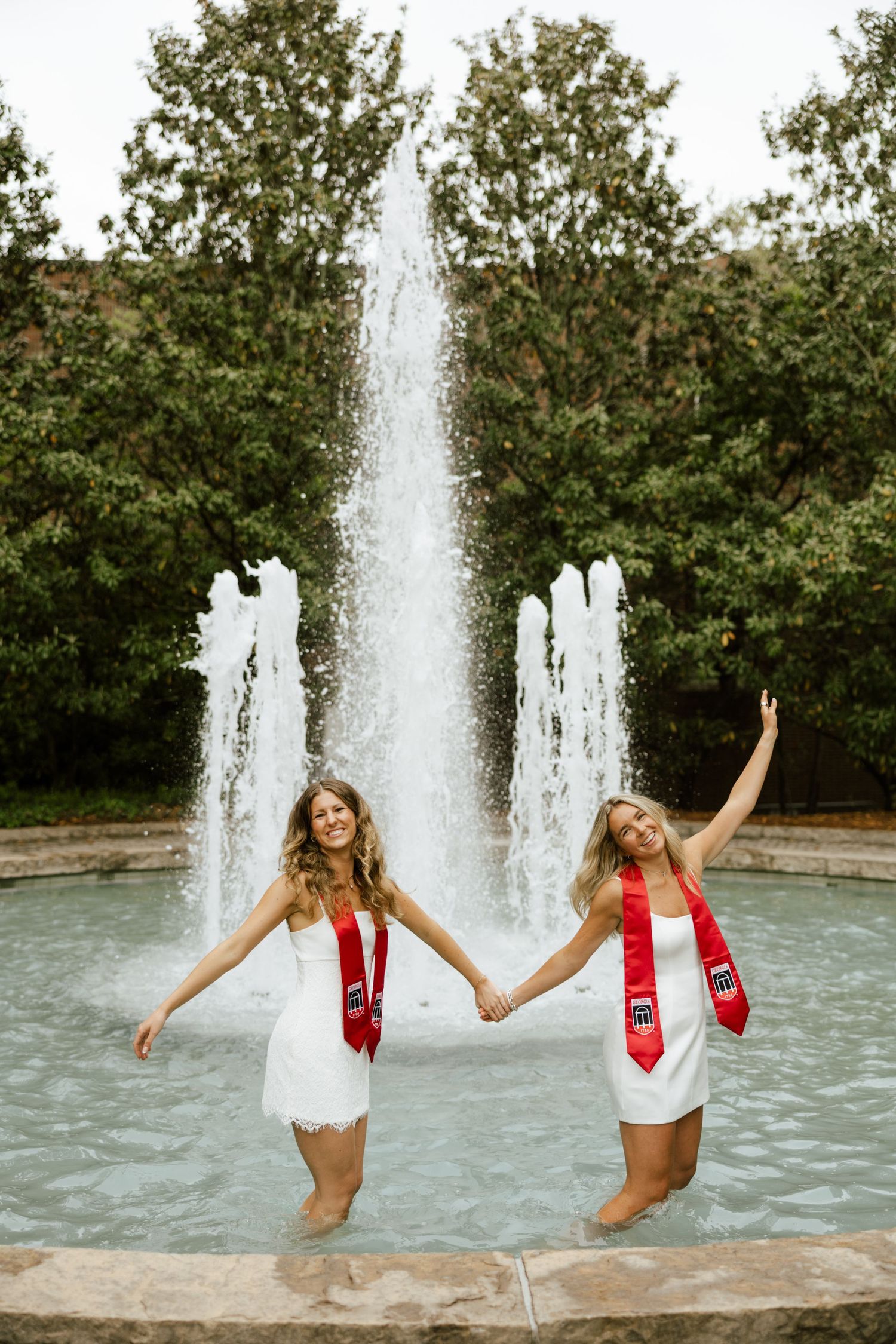 Two graduating women in white dresses and red sashes celebrate in a fountain, holding hands and cheering.