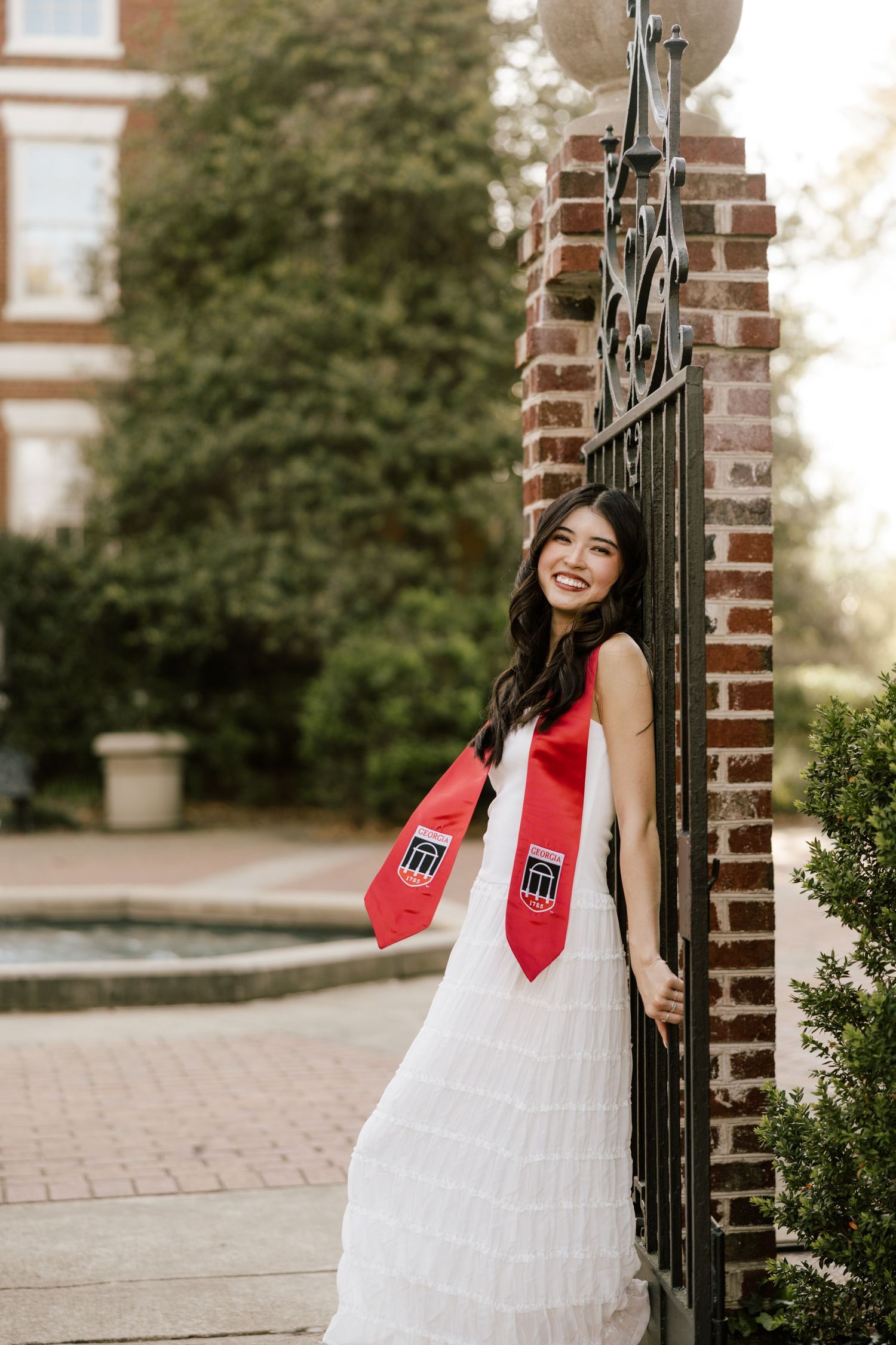 Smiling graduate in white dress and red graduation stole poses by ornate iron gate on brick campus.