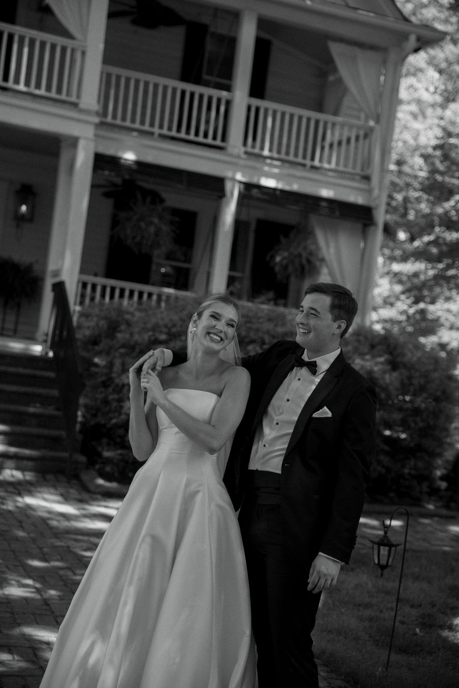 Black and white photo of a laughing bride in a strapless gown and groom in a tuxedo posing outside a historic mansion.