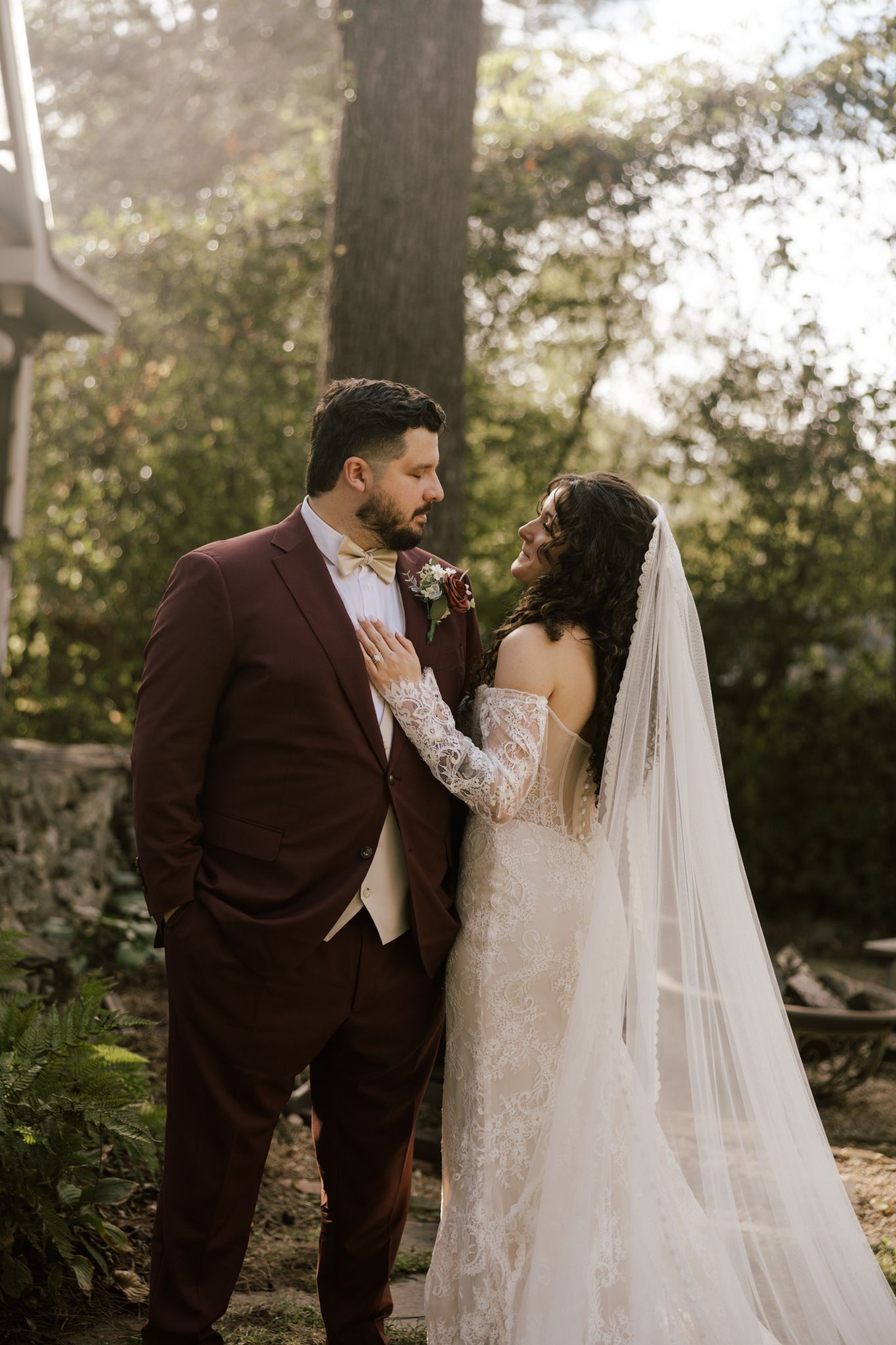 Bride in lace gown with veil and groom in burgundy suit share romantic moment in wooded setting.