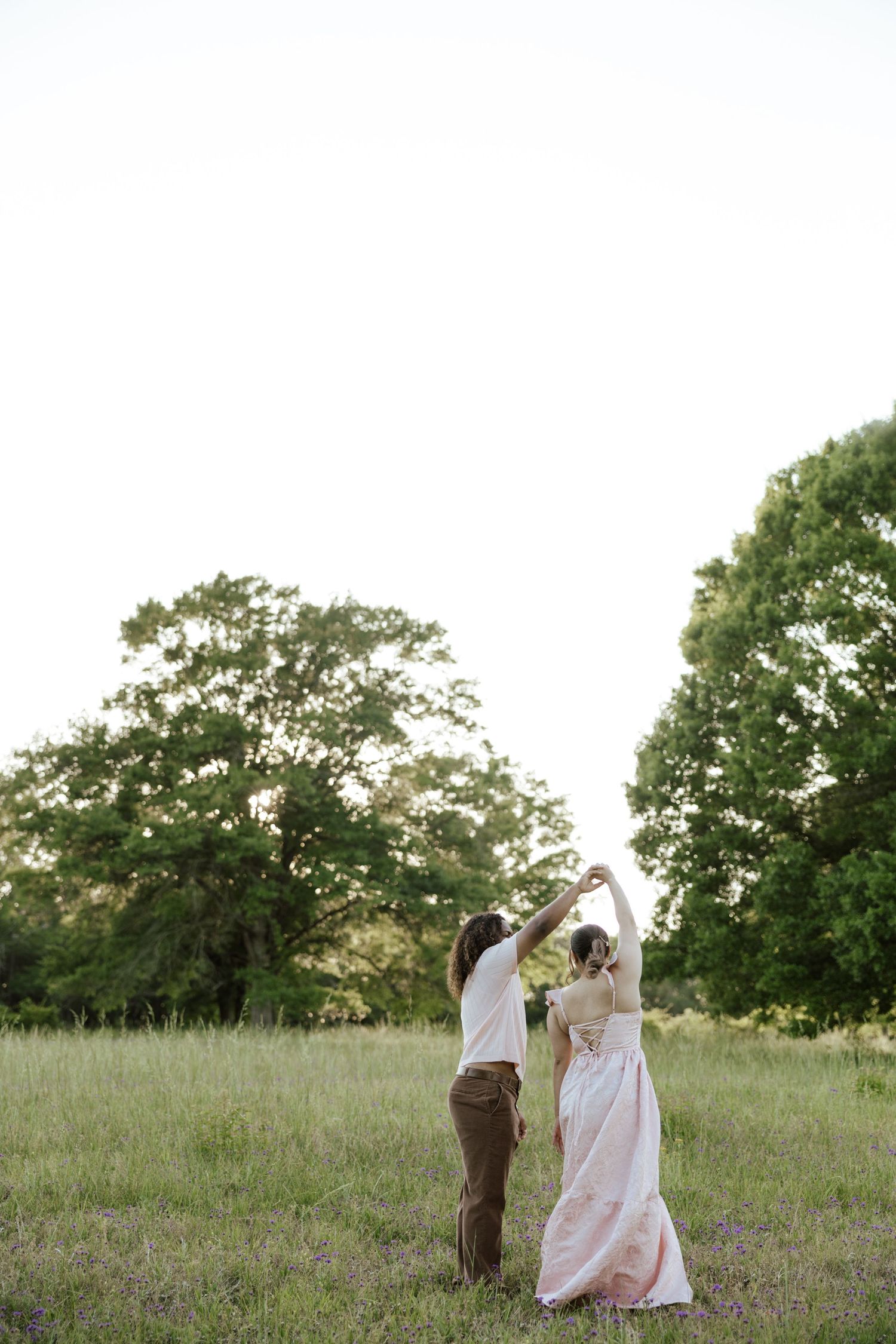 Couple dancing in a green meadow surrounded by trees, wearing elegant attire, in a romantic outdoor setting.