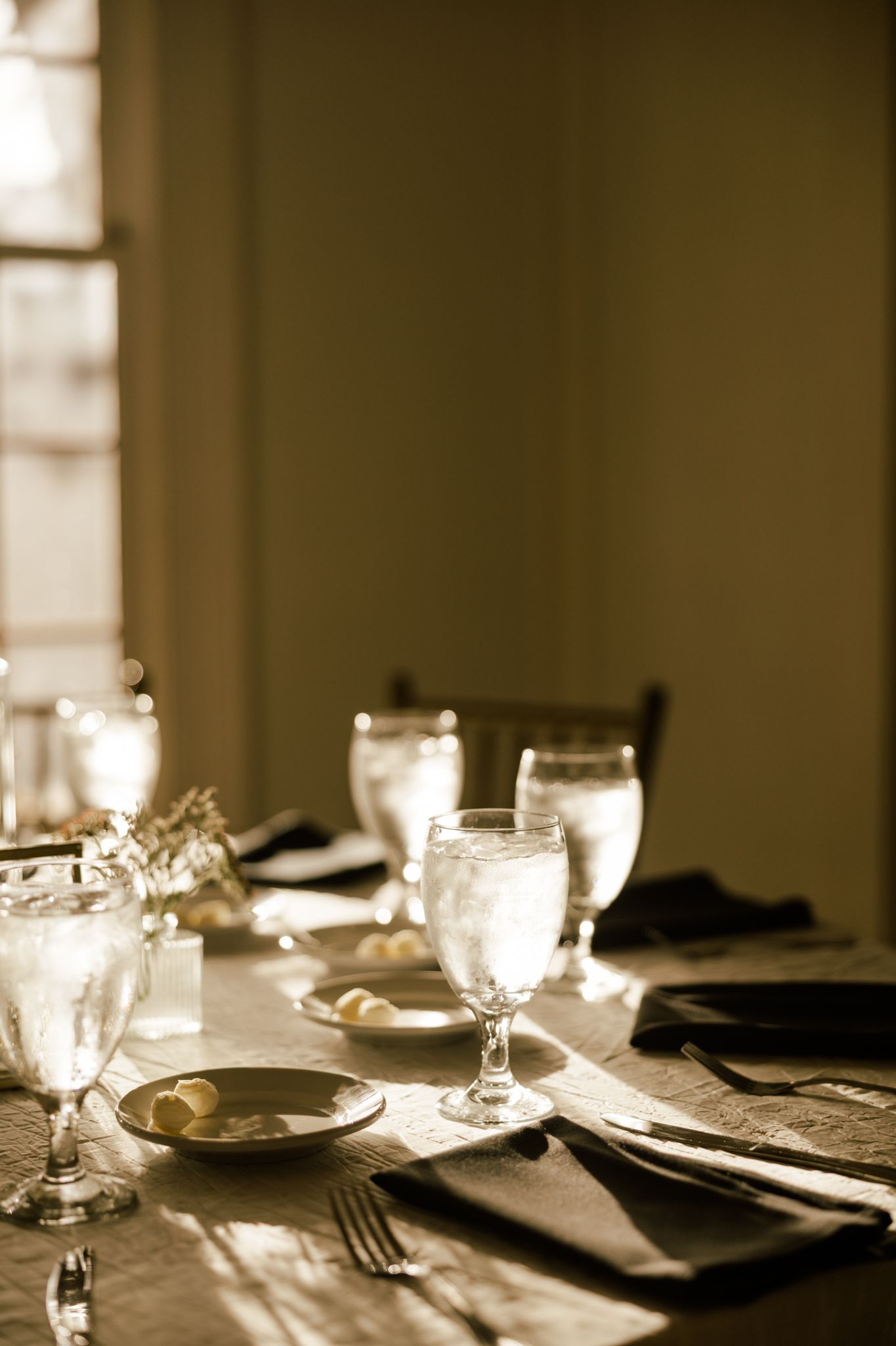 Elegant wedding reception table setting with crystal water glasses and dark linen napkins bathed in warm golden sunlight.