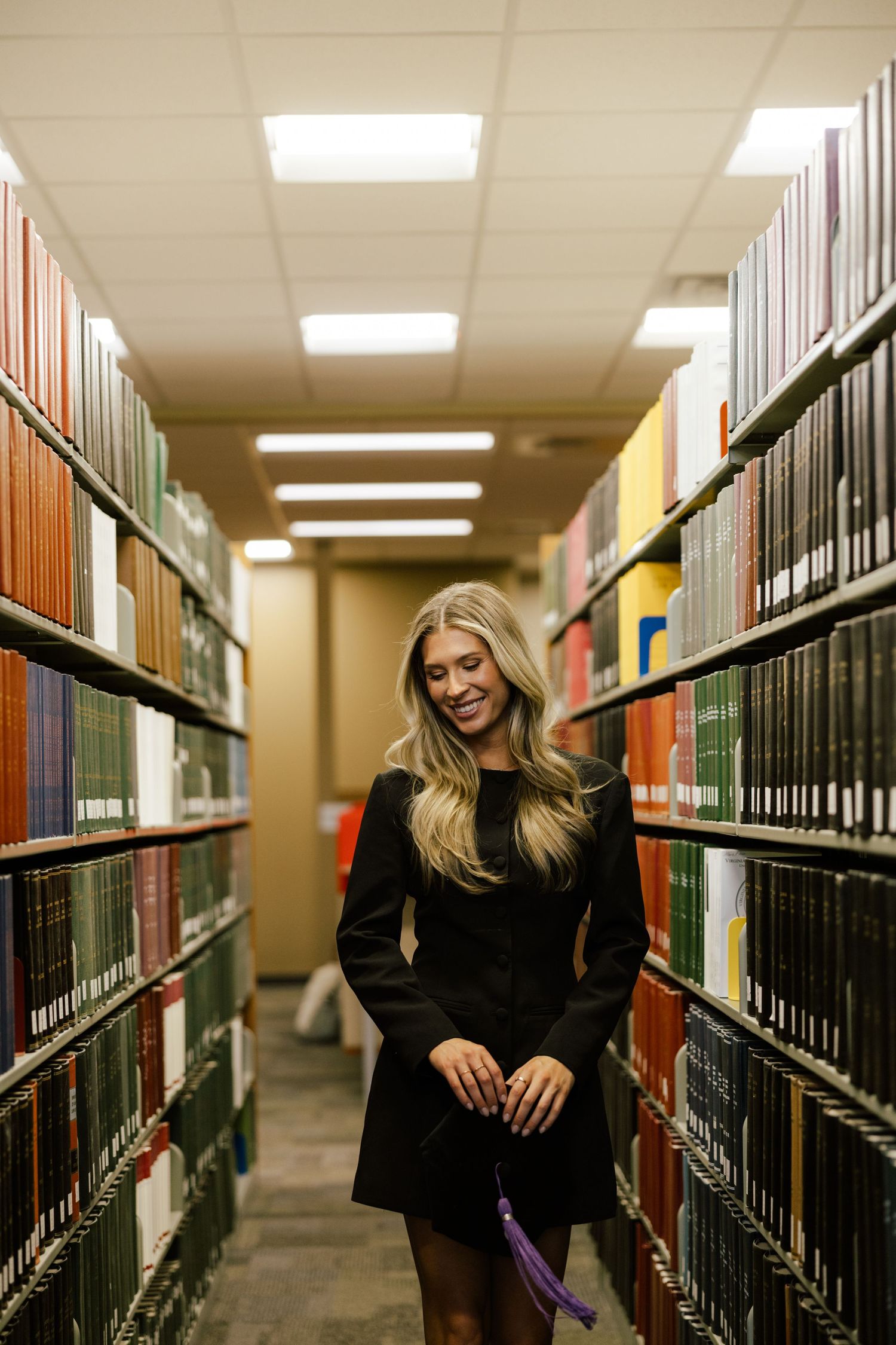 Blonde woman in black dress smiling between library bookshelves holding purple tassel.