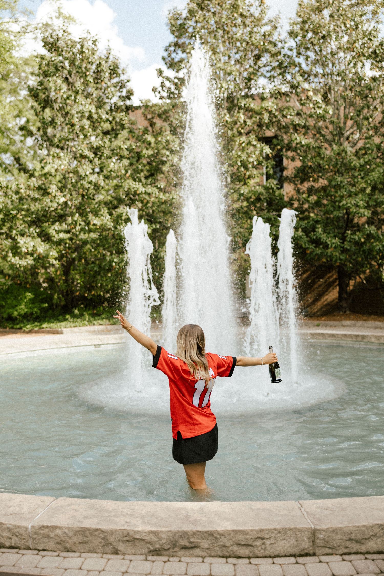 Woman in red jersey wading in fountain with arms outstretched, holding bottle, surrounded by lush green trees.
