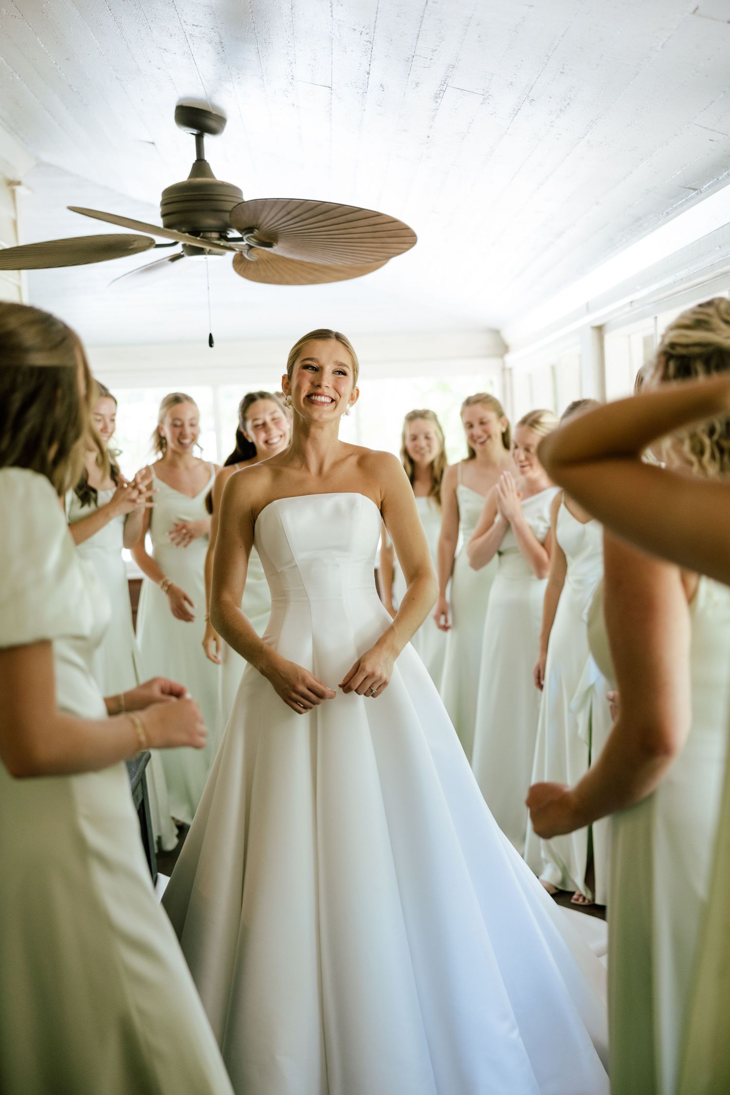 Smiling bride in strapless white gown surrounded by bridesmaids in ivory dresses celebrating in a bright white room.