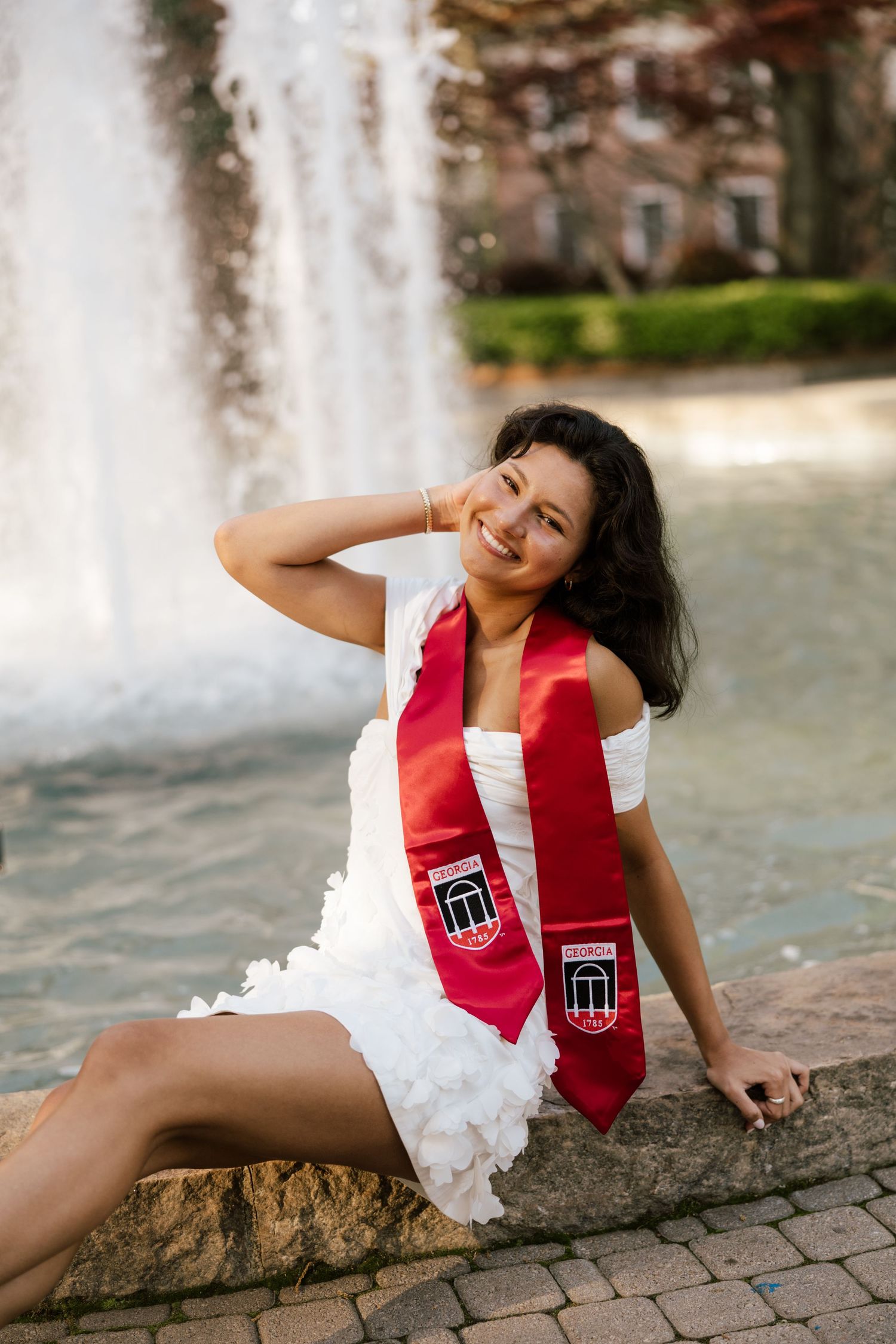 Smiling graduate in white dress and red graduation stole sits by fountain outdoors.