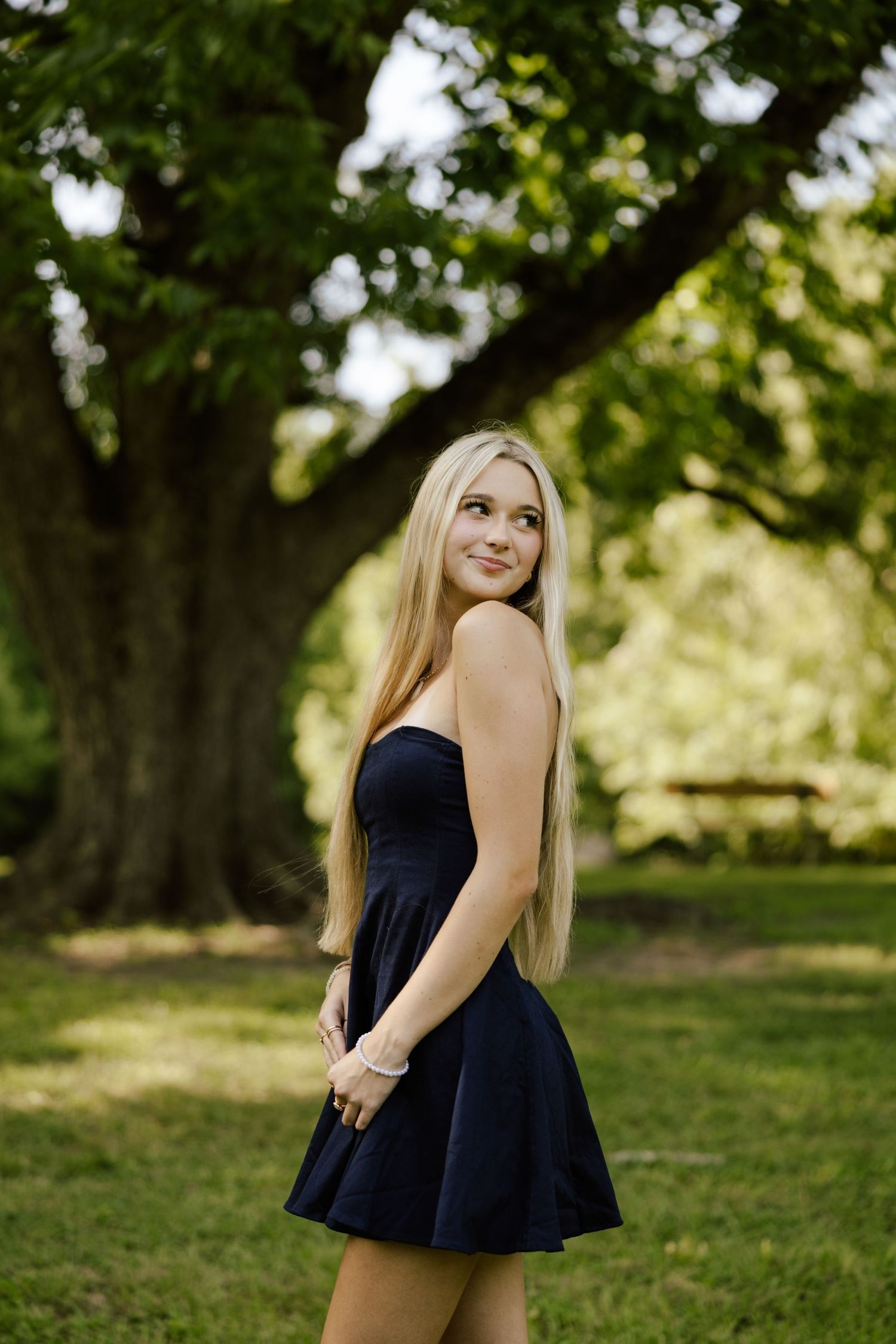 Young blonde woman in a black strapless mini dress smiling outdoors in a lush green park setting.