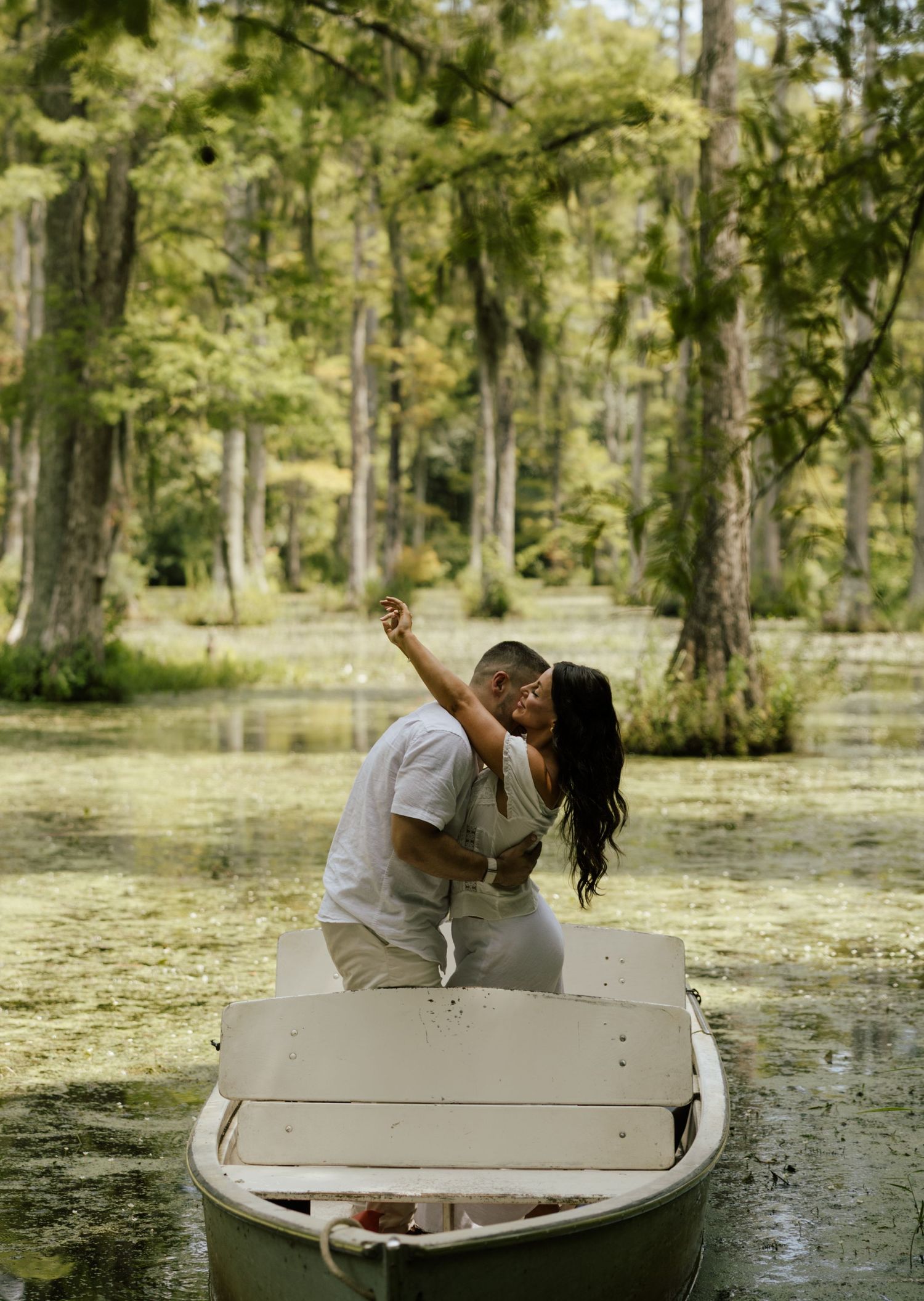 Couple sharing a romantic kiss while taking a selfie in a white rowboat on a serene, moss-covered swamp surrounded by trees.
