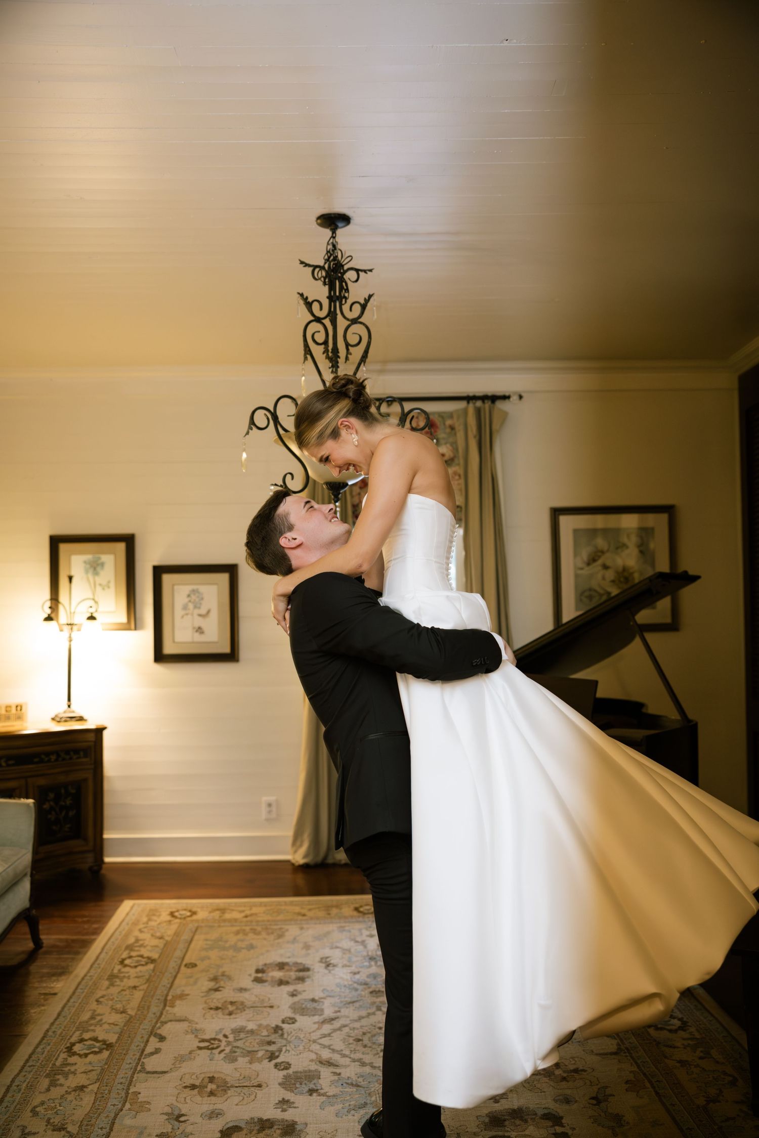 Groom lifts bride in white wedding gown during romantic indoor dance near chandelier and grand piano.