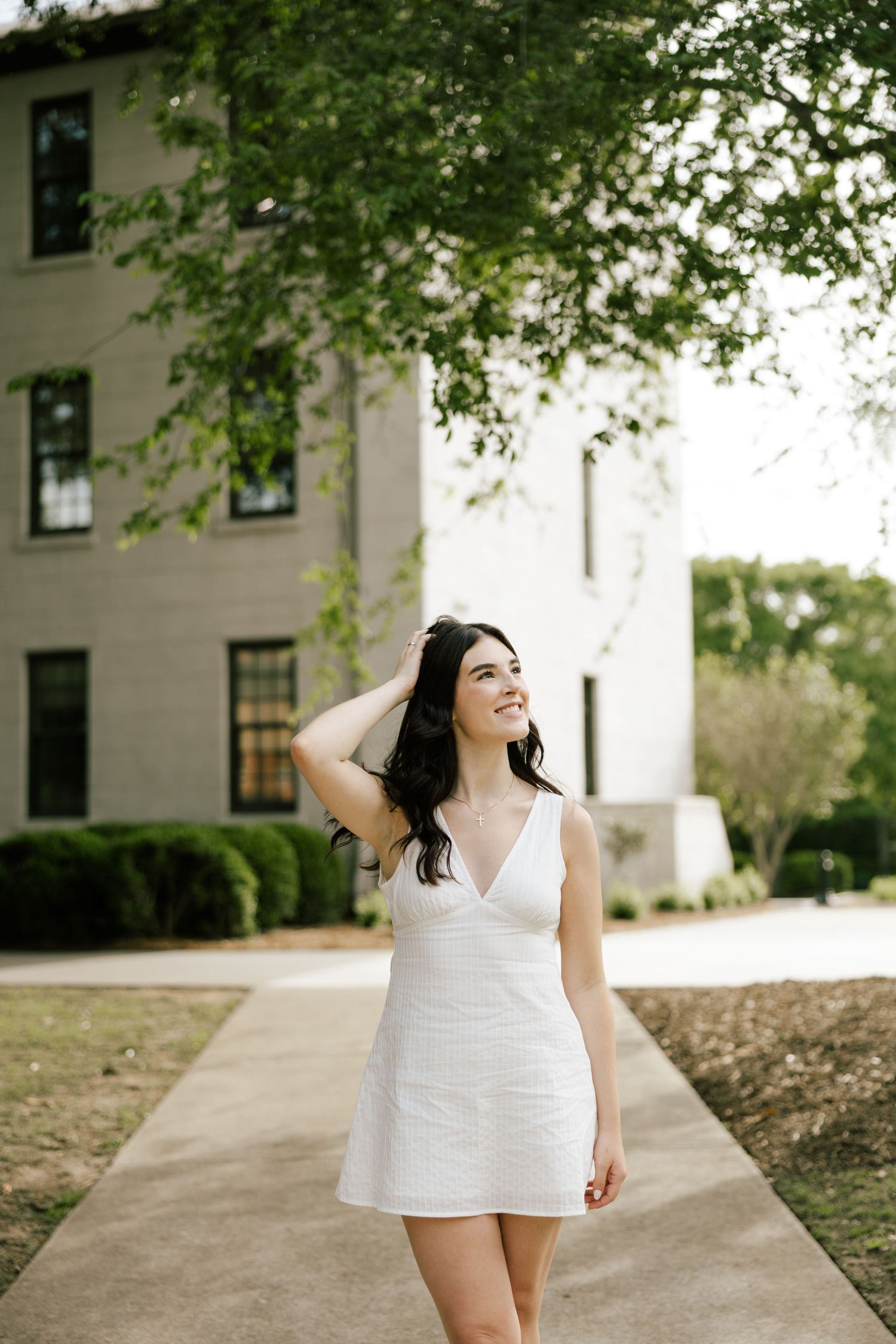A smiling young woman in a white mini dress walks along a sidewalk outside a brick building surrounded by green trees.