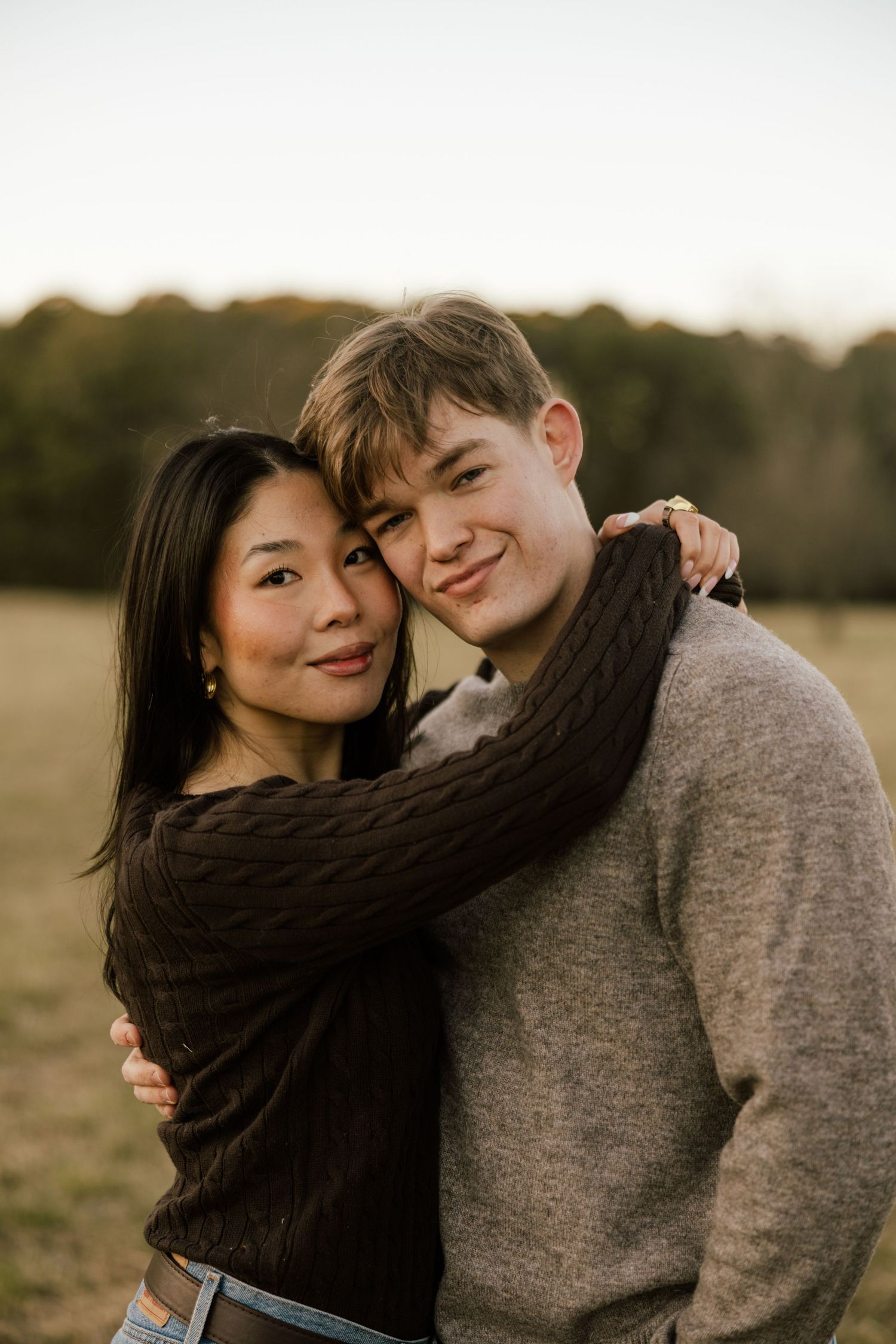 A couple embraces outdoors in a field at sunset, wearing casual sweaters and sharing an intimate moment together.