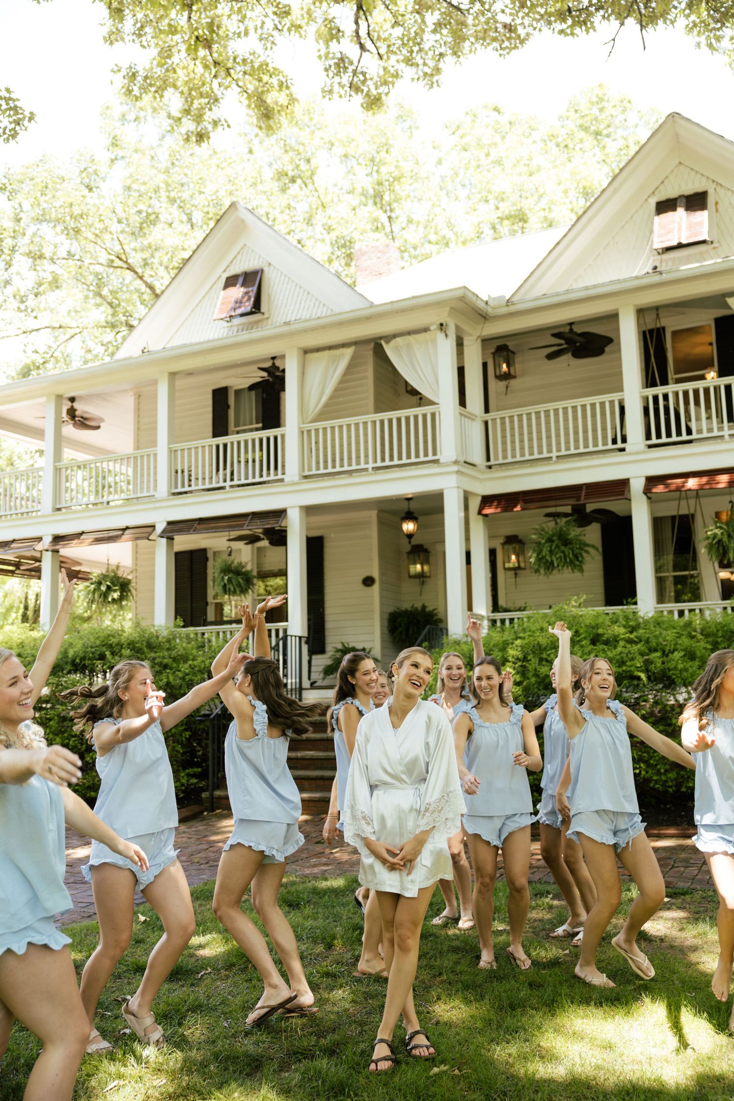 Bride in white robe celebrates with bridesmaids in matching light blue pajamas outside a charming Southern estate.