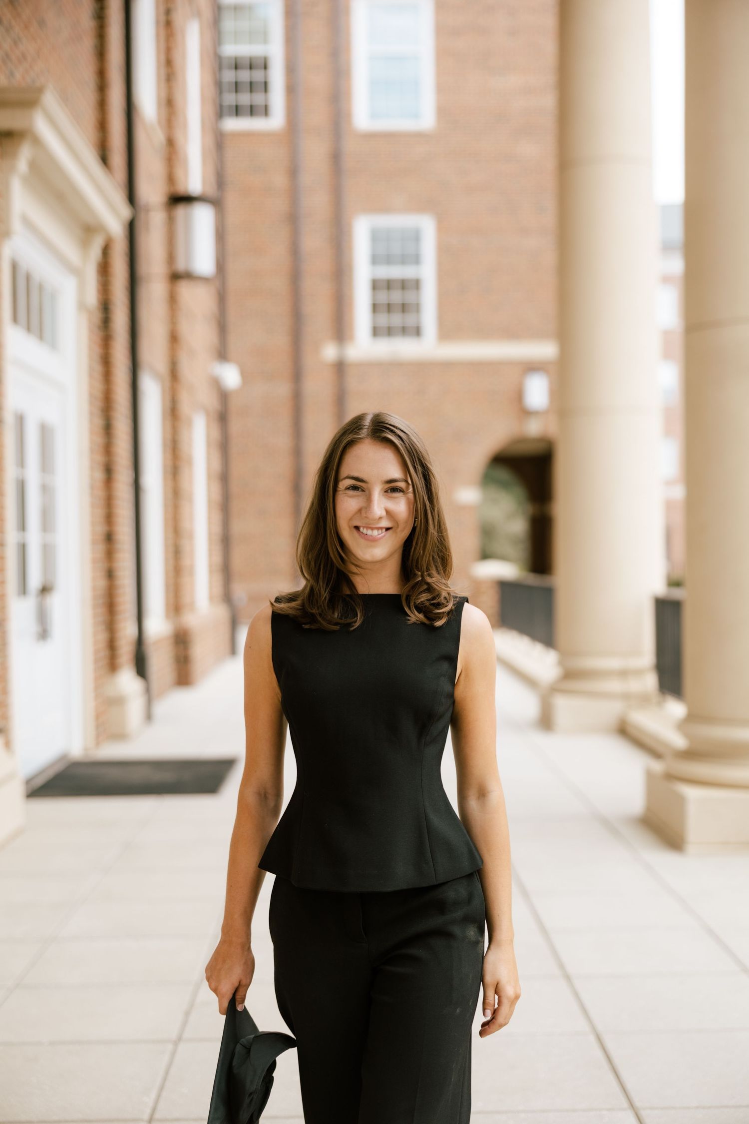 Professional woman in black sleeveless peplum top and trousers smiling outdoors near brick building with columns.