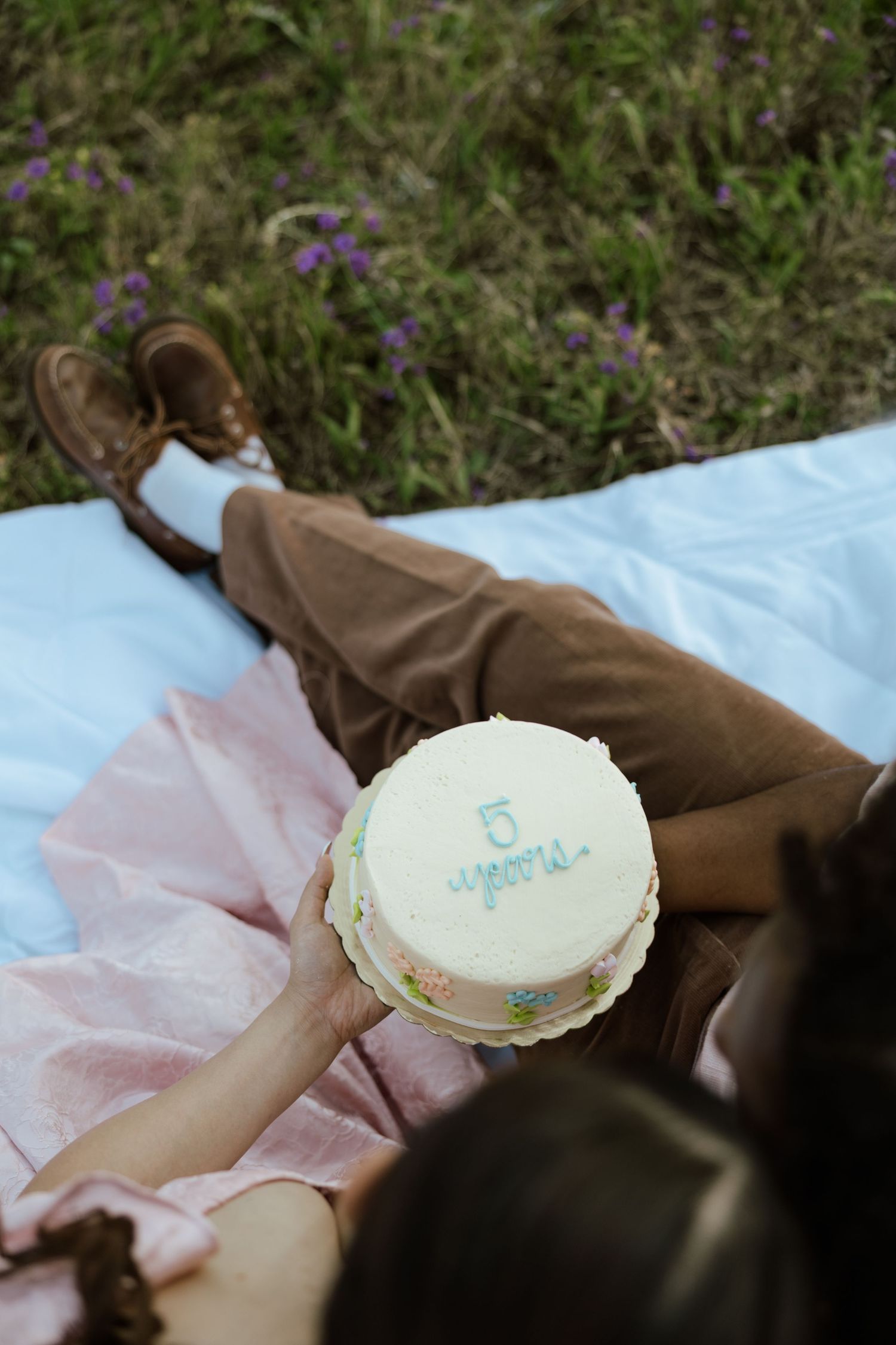 Couple lying on a blanket outdoors holding a white cake reading '5 years' celebrating their anniversary.