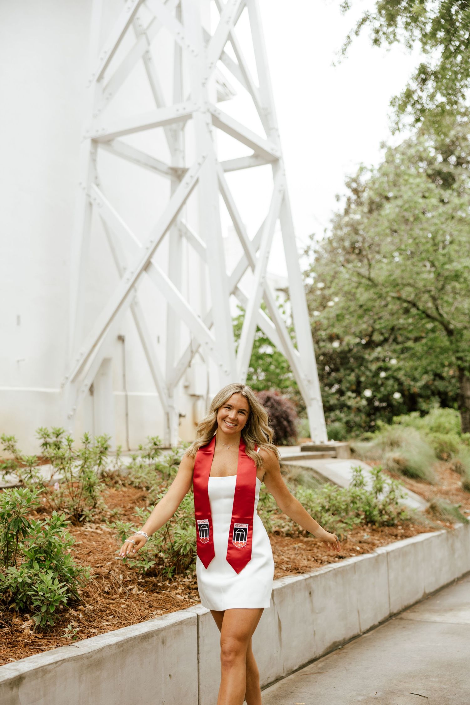 A smiling graduate in a white dress and red sash poses outdoors near a white steel structure and lush greenery.