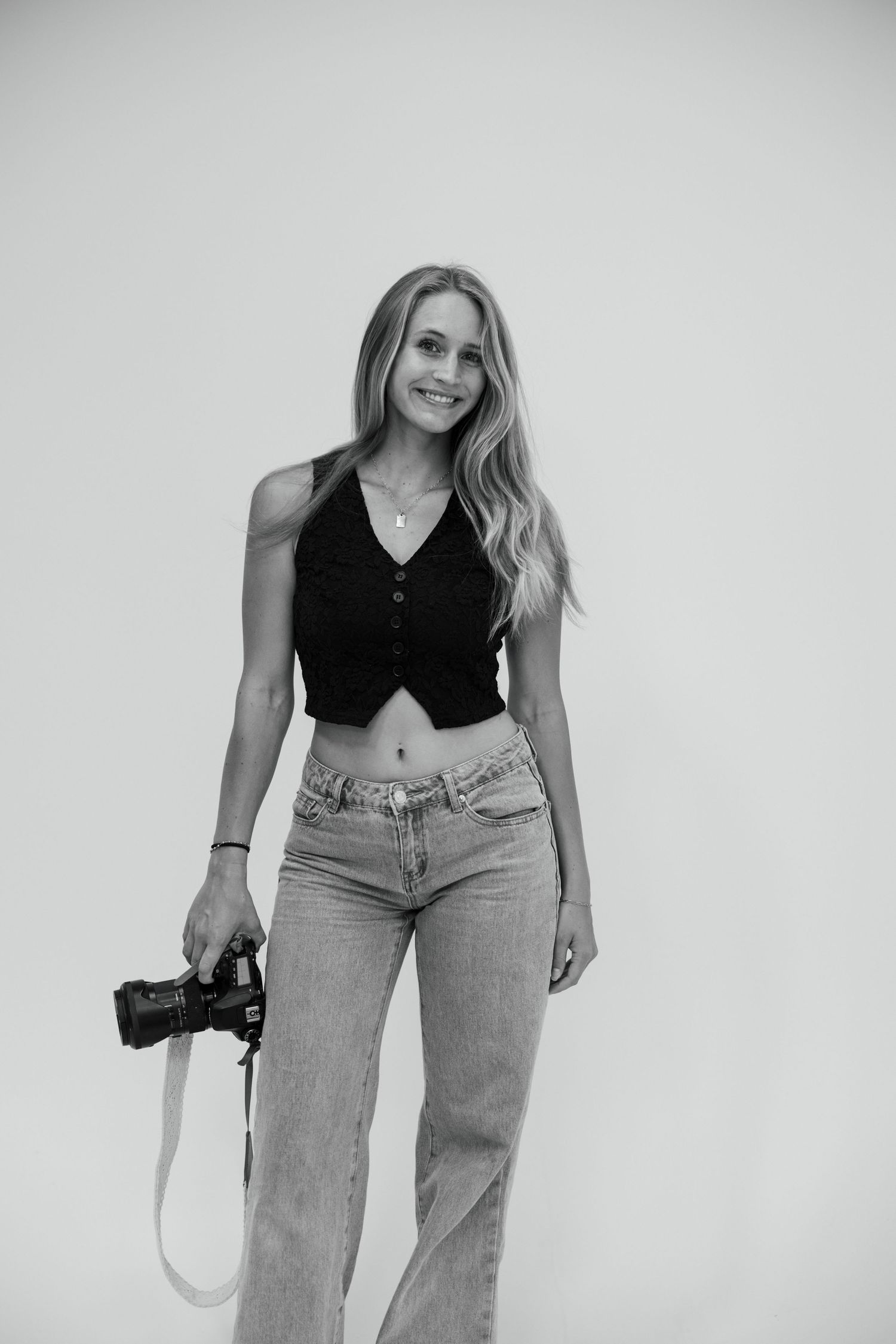Black and white studio portrait of photographer holding camera against plain backdrop.