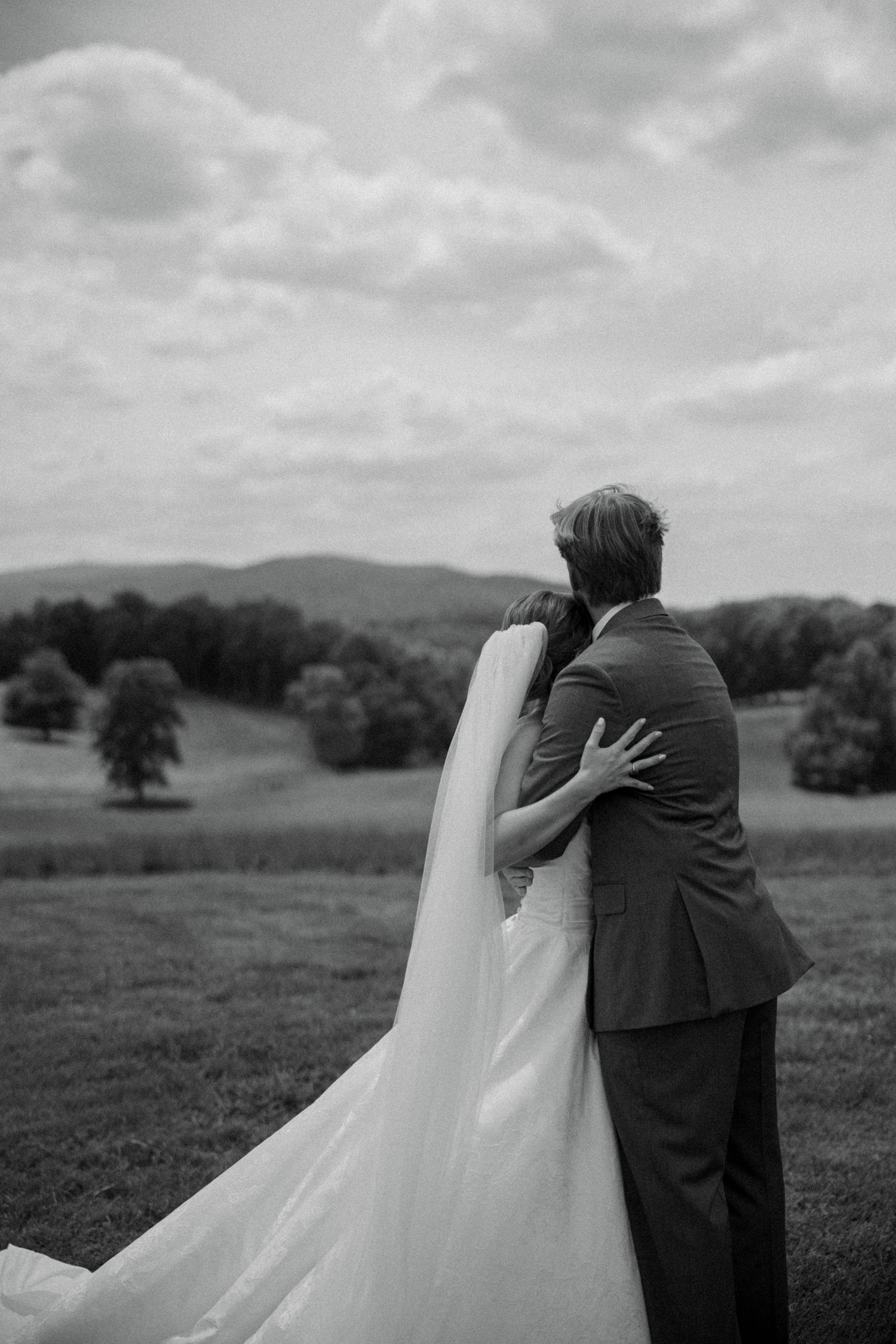 Black and white photo of bride and groom embracing, overlooking rolling hills and trees on their wedding day.