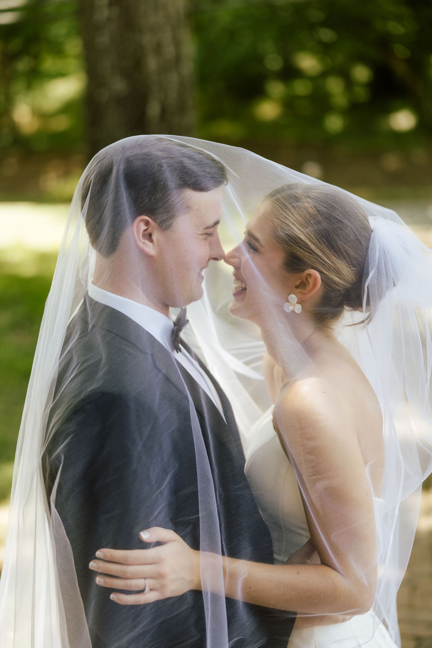 Bride and groom share a romantic moment under a sheer wedding veil outdoors, touching noses and smiling.