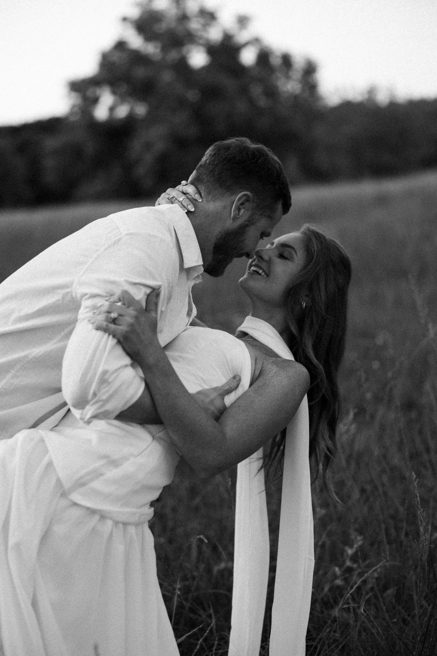 Black and white photo of a couple in white clothing sharing a romantic moment in a lush green field.