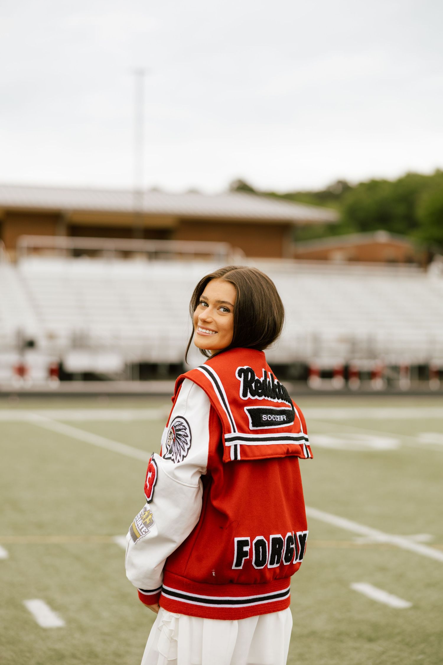 A smiling young woman wearing a red Forsyth letterman jacket poses on a football field, looking back over her shoulder.